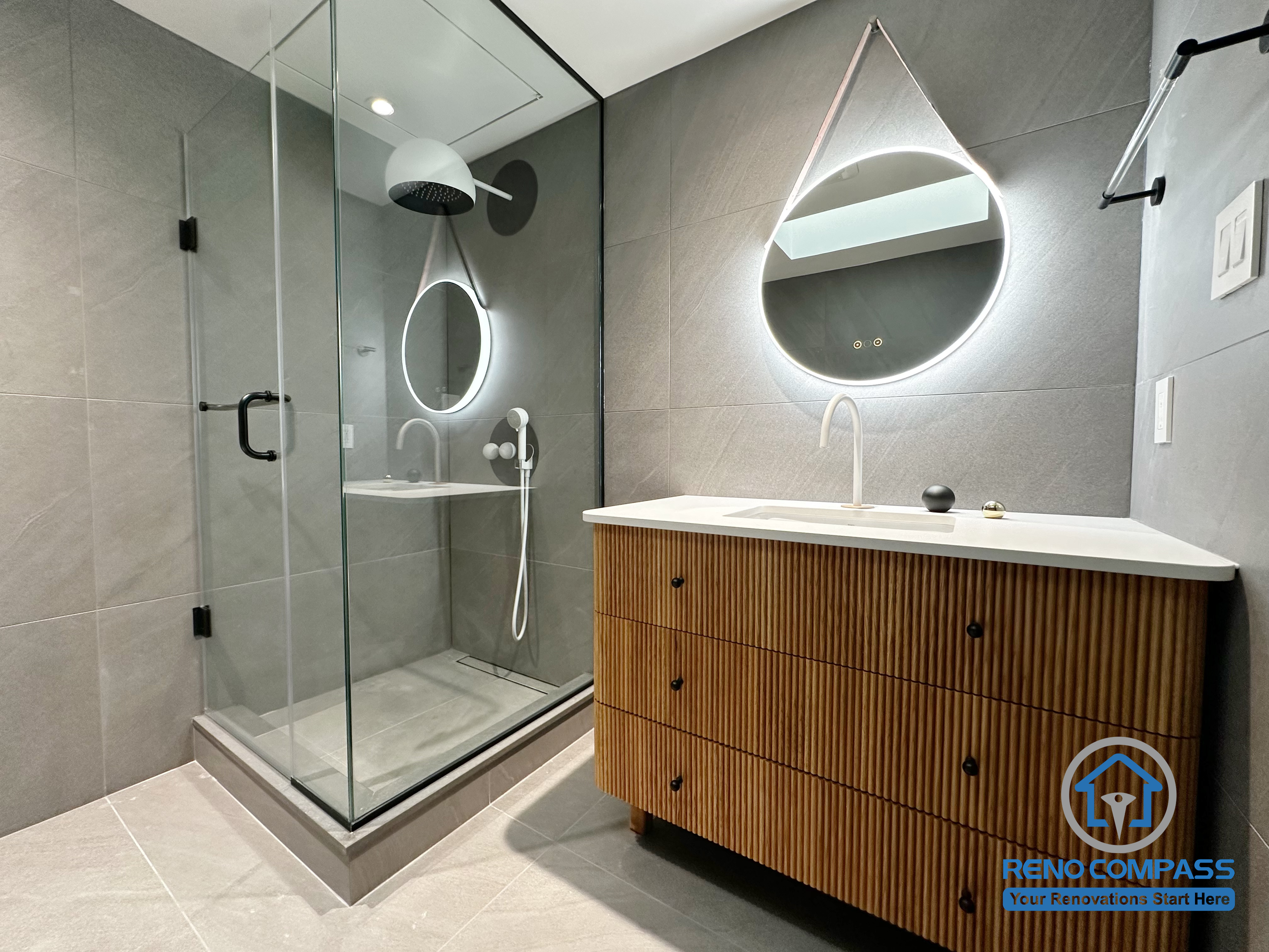 This modern bathroom renovation in Richmond Hill features a frameless glass shower, large-format grey tiles, and a custom wood-textured vanity paired with an integrated sink. Sculptural backlit mirrors and minimalist black fixtures add visual balance, while the clean layout enhances both functionality and comfort. Designed and completed by Reno Compass, this project reflects a refined, contemporary bathroom renovation in the Greater Toronto Area.