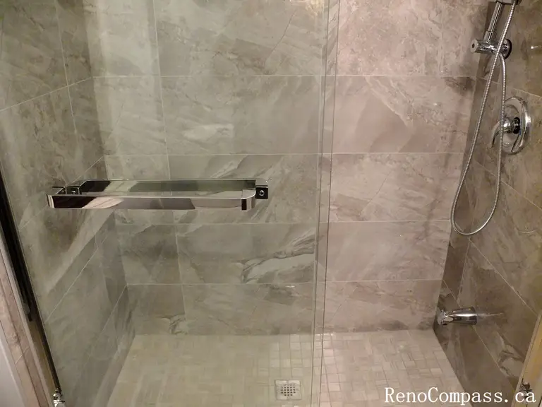 Modern walk-in shower installed during a Richmond Hill interior renovation. The bathroom features marble-look wall tiles, a frameless glass enclosure, a sleek wall-mounted glass bench shelf, and a contemporary handheld shower system. The clean tile layout and minimal hardware create a spacious and elegant shower environment. Completed by Reno Compass Inc., a professional Toronto renovation company specializing in bathroom and interior home renovations.