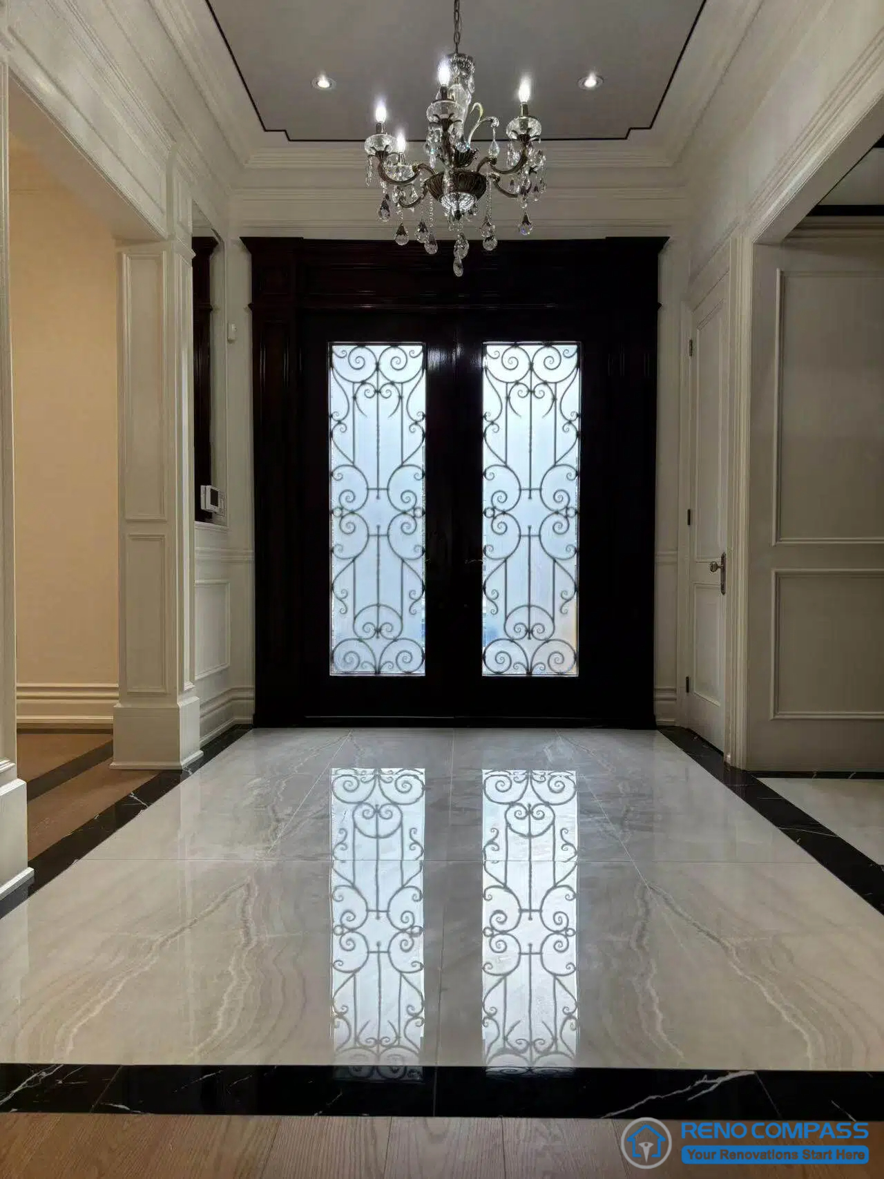 This image presents a European-style entry hall organized along a clear central axis, defined by a decorative double door, marble inlay flooring, and classical wall panelling. The wrought iron detailing on the glass doors introduces refined craftsmanship, while the chandelier and ceiling trim reinforce vertical alignment and proportion. The marble floor pattern establishes a formal arrival sequence, guiding movement into the interior. Together, architectural detailing, material contrast, and symmetry create a composed and ceremonial entrance within a high-end Richmond Hill interior renovation.