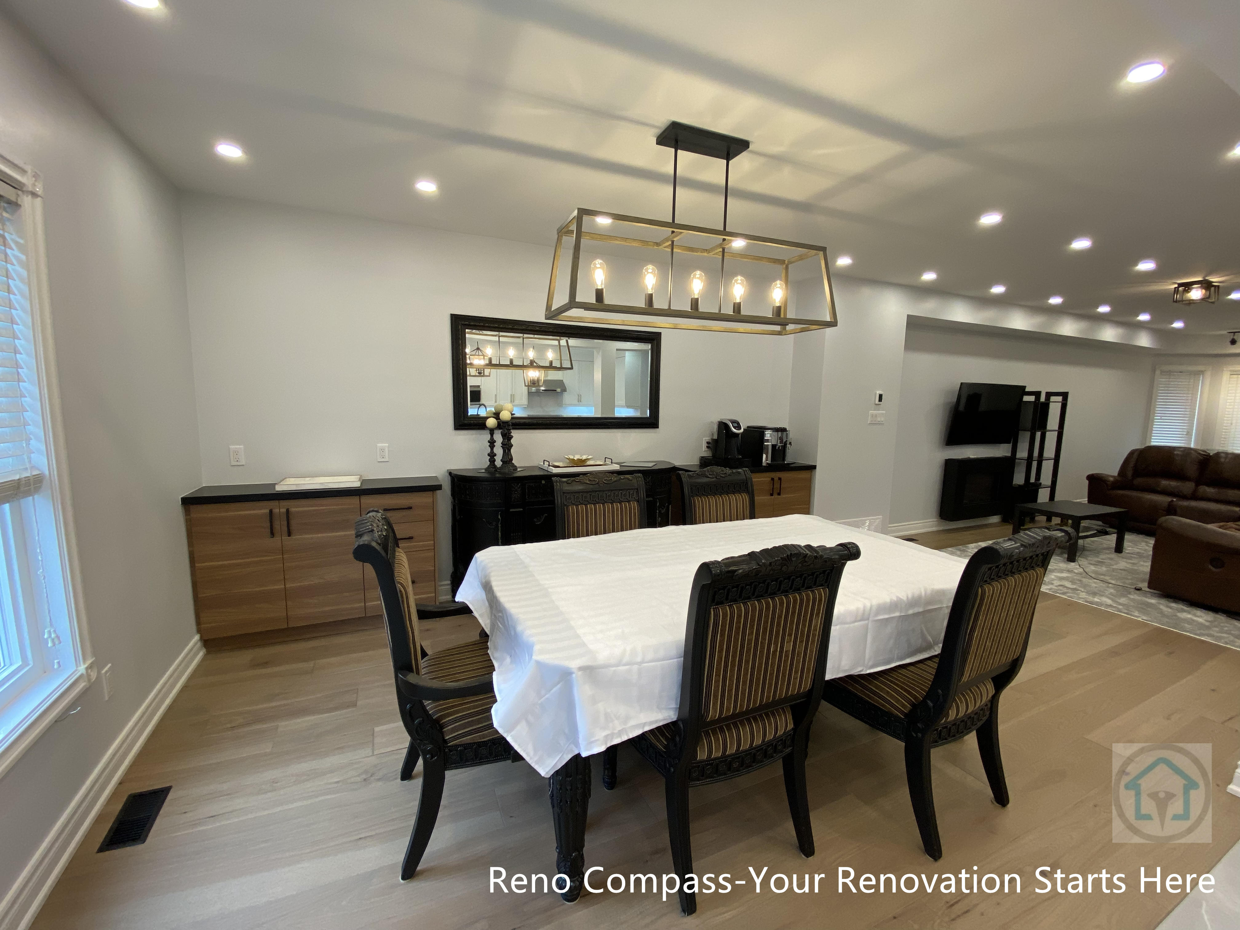 This image showcases a Newmarket interior renovation featuring a spacious open-concept dining area seamlessly connected to the living space. The dining room is anchored by a large rectangular table beneath a modern linear chandelier, creating a clear visual focal point. Recessed ceiling lights provide even illumination throughout the space, enhancing brightness and depth. Warm-toned hardwood flooring extends across the dining and living areas, reinforcing continuity and a cohesive interior flow. Custom side cabinets along the wall add functional storage while maintaining a clean, minimalist aesthetic. Neutral wall colors and balanced lighting contribute to a calm and timeless atmosphere. Completed by a Toronto renovation company, this dining room renovation emphasizes thoughtful layout planning, lighting design, and durable materials suited for everyday family use and entertaining.
