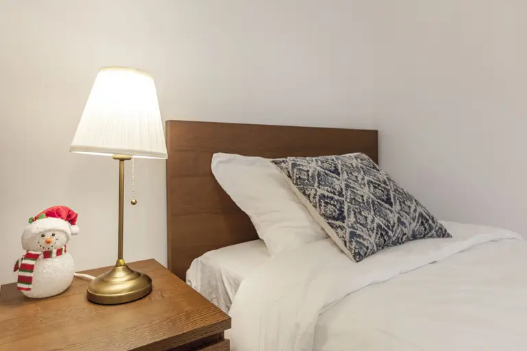 This Toronto bedroom renovation highlights a warm and cozy sleeping environment with a clean bed setup, wooden headboard, and matching nightstand. Soft lighting from the bedside lamp enhances comfort, creating a relaxing atmosphere ideal for modern living. The simple yet functional design reflects the attention to detail of a professional Toronto Renovation Company.