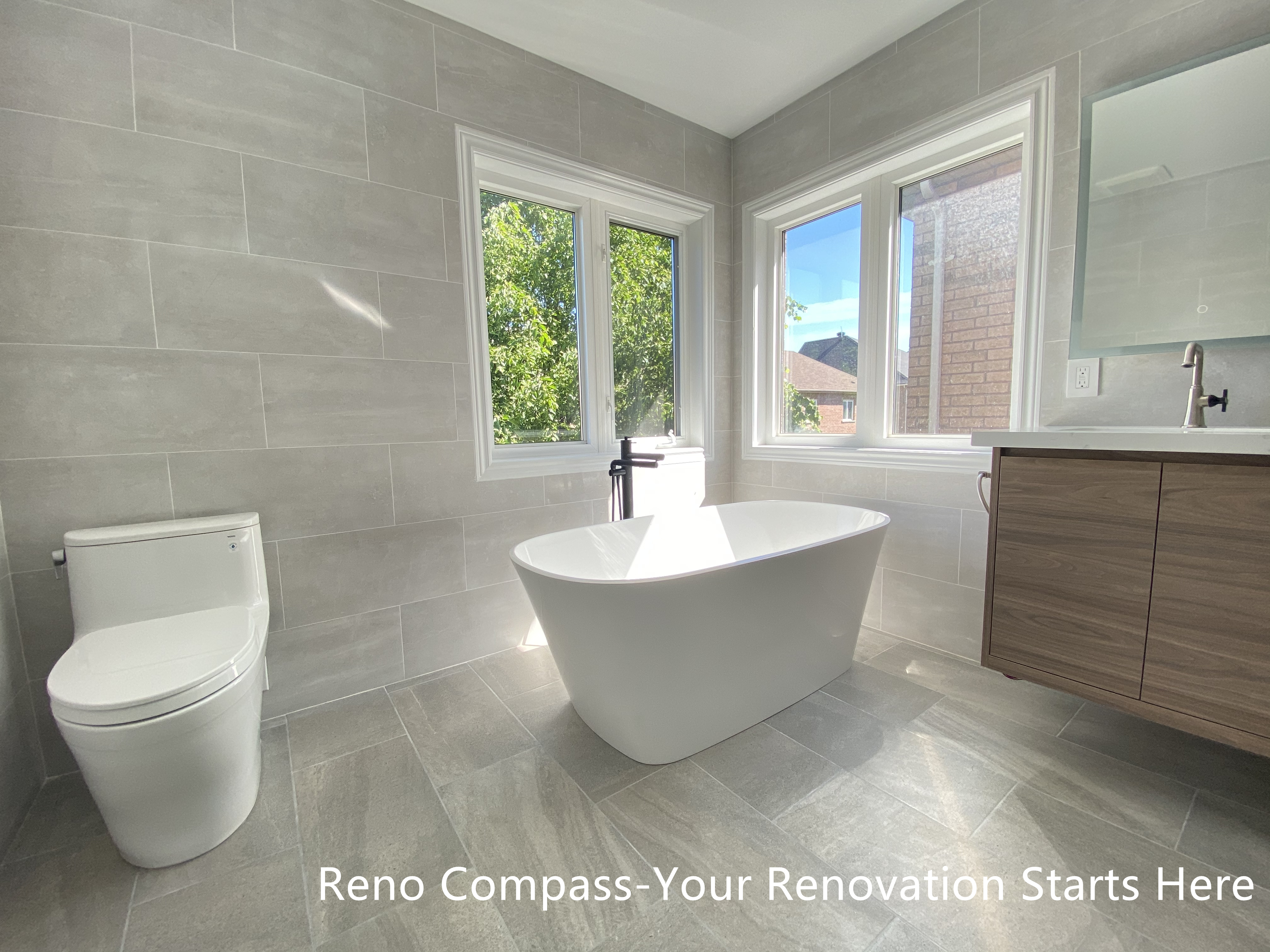 This Aurora bathroom renovation highlights a freestanding bathtub positioned beside corner windows, allowing natural light to fill the space throughout the day. Large-format tile flooring and wall finishes create a seamless, modern backdrop, while the clean tub silhouette and minimalist fixtures reinforce a calm, spa-inspired atmosphere. The layout maintains generous circulation and visual openness, making the bathing area both functional and relaxing. Designed and completed by Reno Compass, this bathroom renovation emphasizes comfort, proportion, and long-term usability.