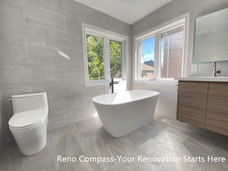This Aurora bathroom renovation highlights a freestanding bathtub positioned beside corner windows, allowing natural light to fill the space throughout the day. Large-format tile flooring and wall finishes create a seamless, modern backdrop, while the clean tub silhouette and minimalist fixtures reinforce a calm, spa-inspired atmosphere. The layout maintains generous circulation and visual openness, making the bathing area both functional and relaxing. Designed and completed by Reno Compass, this bathroom renovation emphasizes comfort, proportion, and long-term usability.