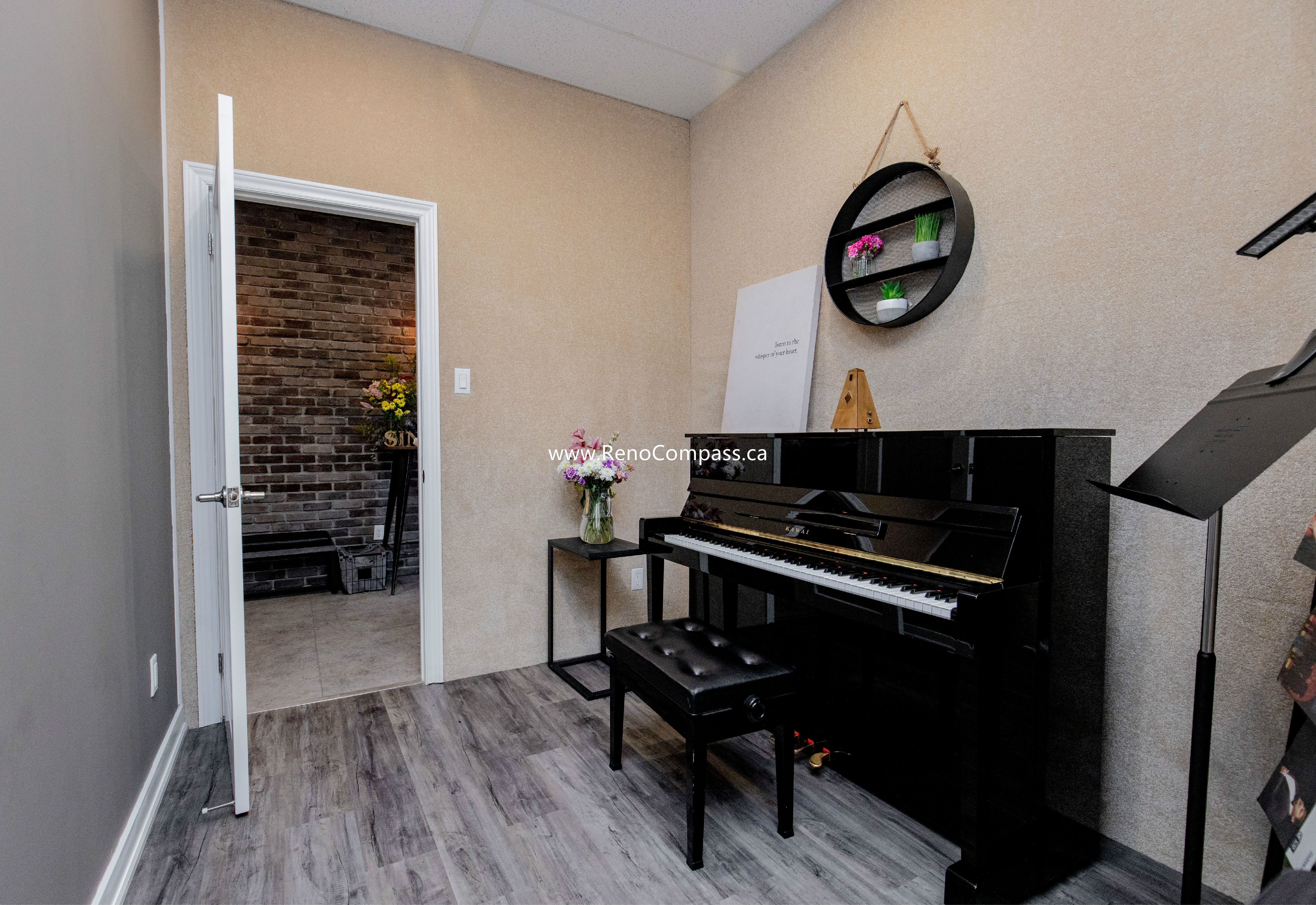 A cozy and professional private lesson room at The Singing School. This commercial interior features modern grey wood laminate flooring, custom textured wall finishes, and a glimpse into a feature room with industrial brick-style wallpaper. Expert commercial contracting in Richmond Hill.