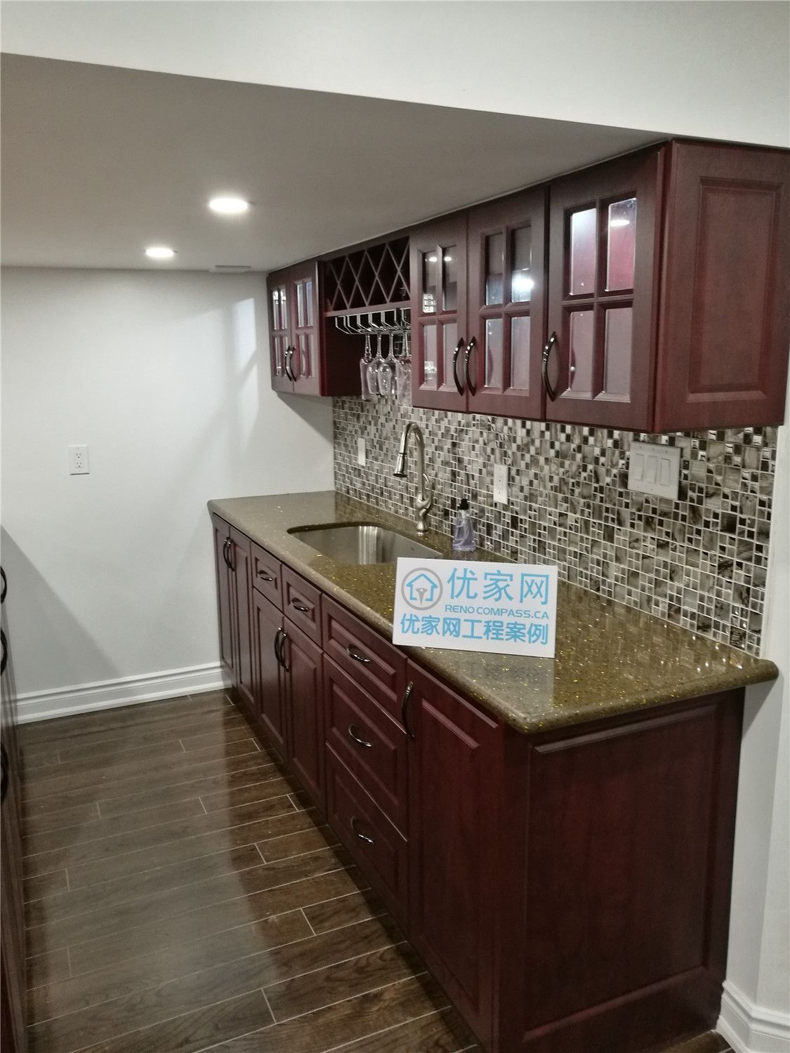 This Markham basement renovation highlights a custom-built wet bar featuring rich cherry-toned cabinetry, a granite countertop, and a detailed mosaic tile backsplash. Upper glass-front cabinets and a built-in wine rack add both functionality and visual appeal, creating the perfect entertainment corner for hosting guests. Recessed lighting enhances the warmth of the wood finishes while maintaining a bright, modern atmosphere. Professionally designed and installed, this basement bar upgrade combines practicality with upscale craftsmanship, elevating the overall basement living experience in Markham.