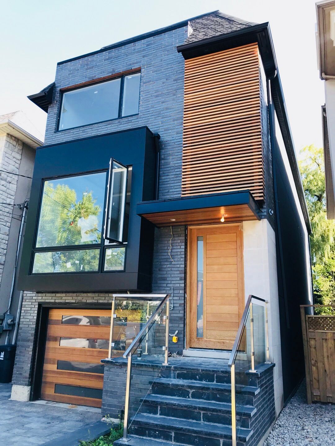 A stunning Toronto modern style renovation featuring dark charcoal brick, a black metal cantilevered window box, and natural cedar slat accents. This narrow-lot custom home maximizes curb appeal with a contemporary glass railing and matching wood garage door.