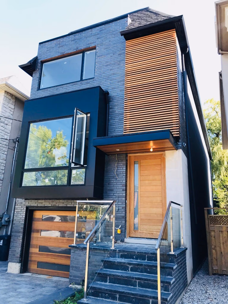A stunning Toronto modern style renovation featuring dark charcoal brick, a black metal cantilevered window box, and natural cedar slat accents. This narrow-lot custom home maximizes curb appeal with a contemporary glass railing and matching wood garage door.