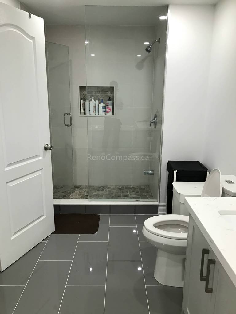 This Toronto bathroom renovation by Reno Compass features a sleek walk-in glass shower with a built-in wall niche for convenient storage and a clean, modern look. Large-format wall tiles and dark floor tiles create a stylish contrast while improving durability and easy maintenance. The upgraded vanity, toilet, and lighting enhance functionality and comfort, delivering a contemporary bathroom space designed for everyday use and long-term value.