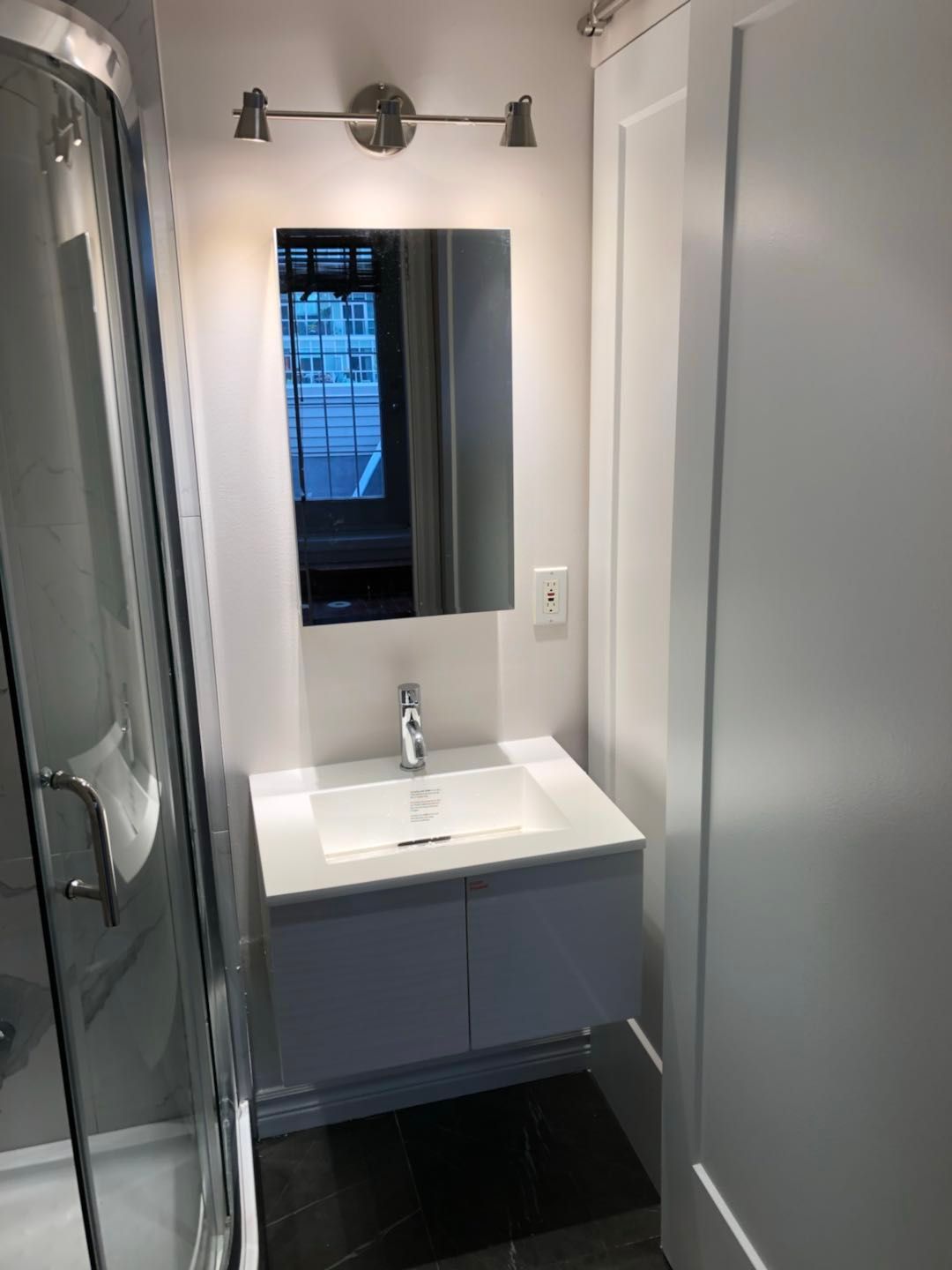This Toronto bathroom renovation highlights a compact yet functional design featuring a modern floating vanity, sleek mirror with wall lighting, and glass shower enclosure. The dark floor tiles and clean white finishes create a contemporary contrast, maximizing both style and practicality. The upgraded layout improves storage, lighting, and everyday comfort, making the small bathroom feel bright, efficient, and modern.
