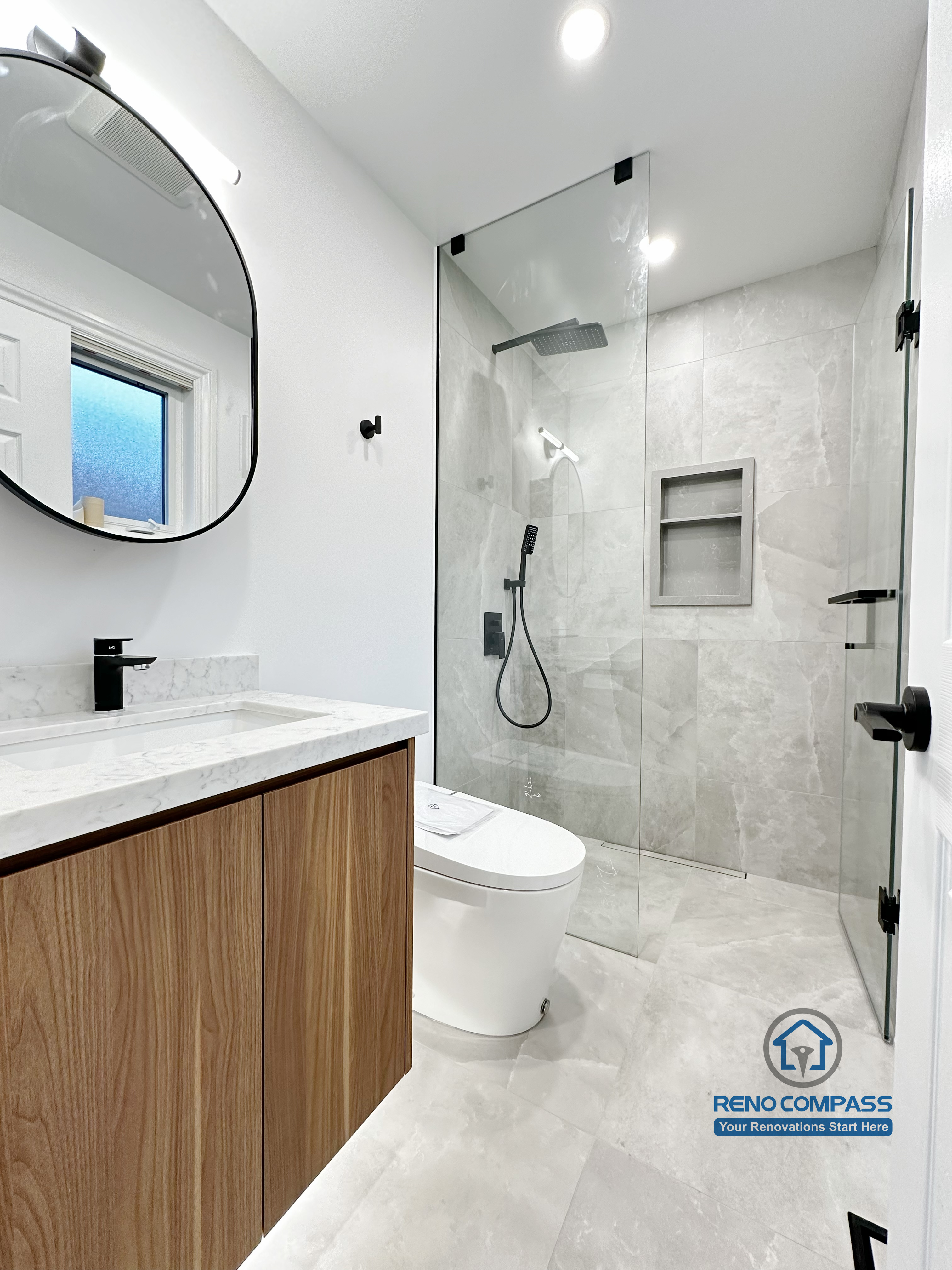 Bathroom renovation in Markham featuring a custom wood vanity with quartz countertop, round wall-mounted mirror, and a frameless glass shower with built-in niche. Clean lines, neutral finishes, and modern fixtures create a bright and functional bathroom layout.
