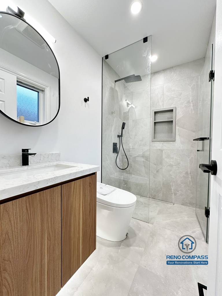 Bathroom renovation in Markham featuring a custom wood vanity with quartz countertop, round wall-mounted mirror, and a frameless glass shower with built-in niche. Clean lines, neutral finishes, and modern fixtures create a bright and functional bathroom layout.