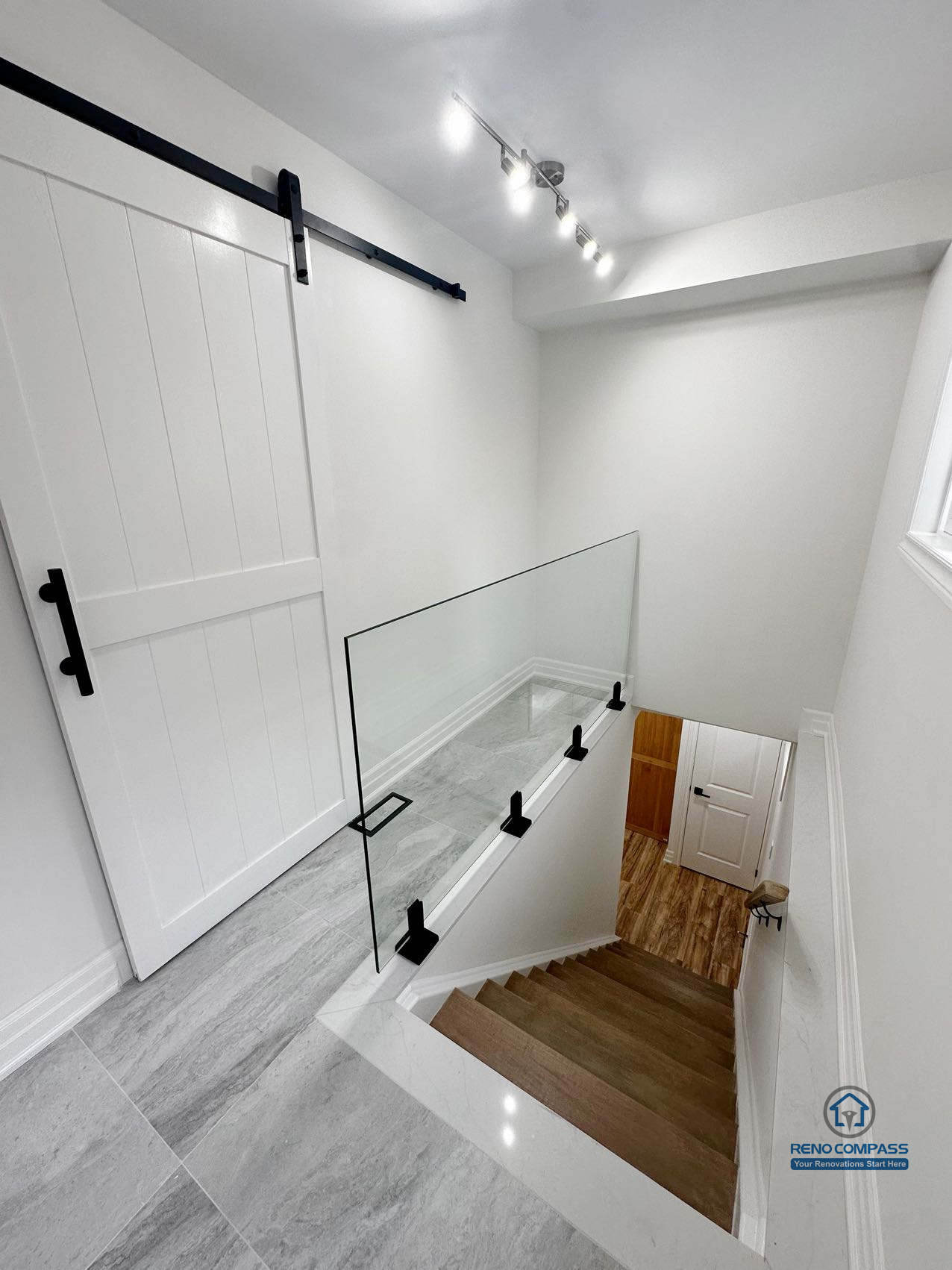This image highlights the upper stair landing of a completed basement renovation in Pickering, featuring a glass guardrail that enhances safety while maintaining visual openness. The stair landing is finished with durable tile flooring, creating a clear transition between levels. A sliding barn-style door provides flexible room separation without interrupting circulation, while track lighting improves visibility and usability in this transitional space. Clean wall finishes and consistent material choices help the stair area integrate seamlessly with the surrounding basement layout.
