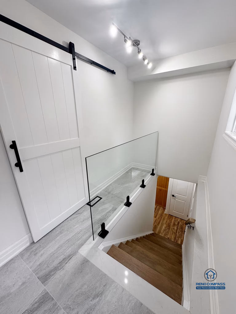 This image highlights the upper stair landing of a completed basement renovation in Pickering, featuring a glass guardrail that enhances safety while maintaining visual openness. The stair landing is finished with durable tile flooring, creating a clear transition between levels. A sliding barn-style door provides flexible room separation without interrupting circulation, while track lighting improves visibility and usability in this transitional space. Clean wall finishes and consistent material choices help the stair area integrate seamlessly with the surrounding basement layout.