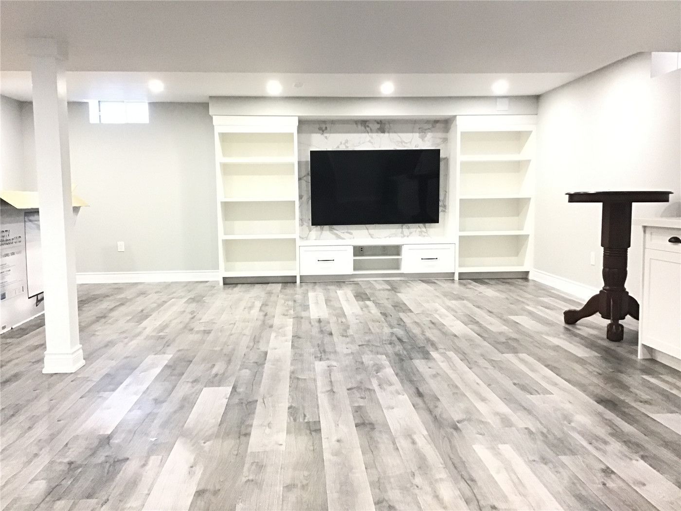 Spacious open-concept basement renovation in Newmarket featuring custom built-in shelving, a modern TV feature wall with marble-style backdrop, and sleek grey vinyl flooring. This functional and elegant design creates the perfect entertainment and family living space.