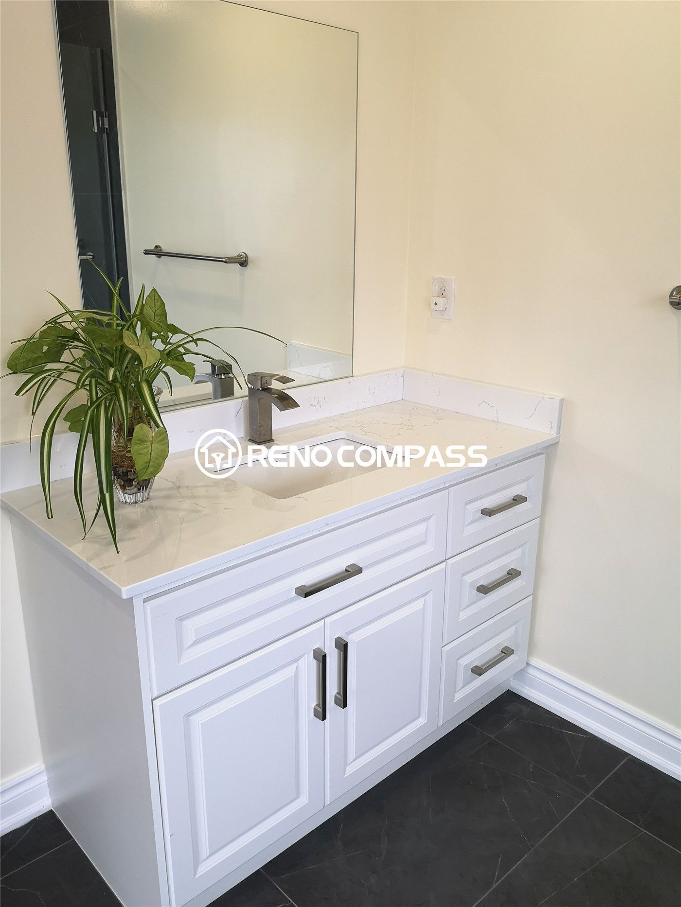 This Toronto bathroom renovation features a crisp white vanity with brushed nickel hardware and a sleek quartz countertop. The minimalist design is complemented by a dark tile floor, creating a striking contrast that enhances the room’s modern elegance. Finished with plant decor for a refreshing touch, this Reno Compass project combines functionality and style in perfect balance.
