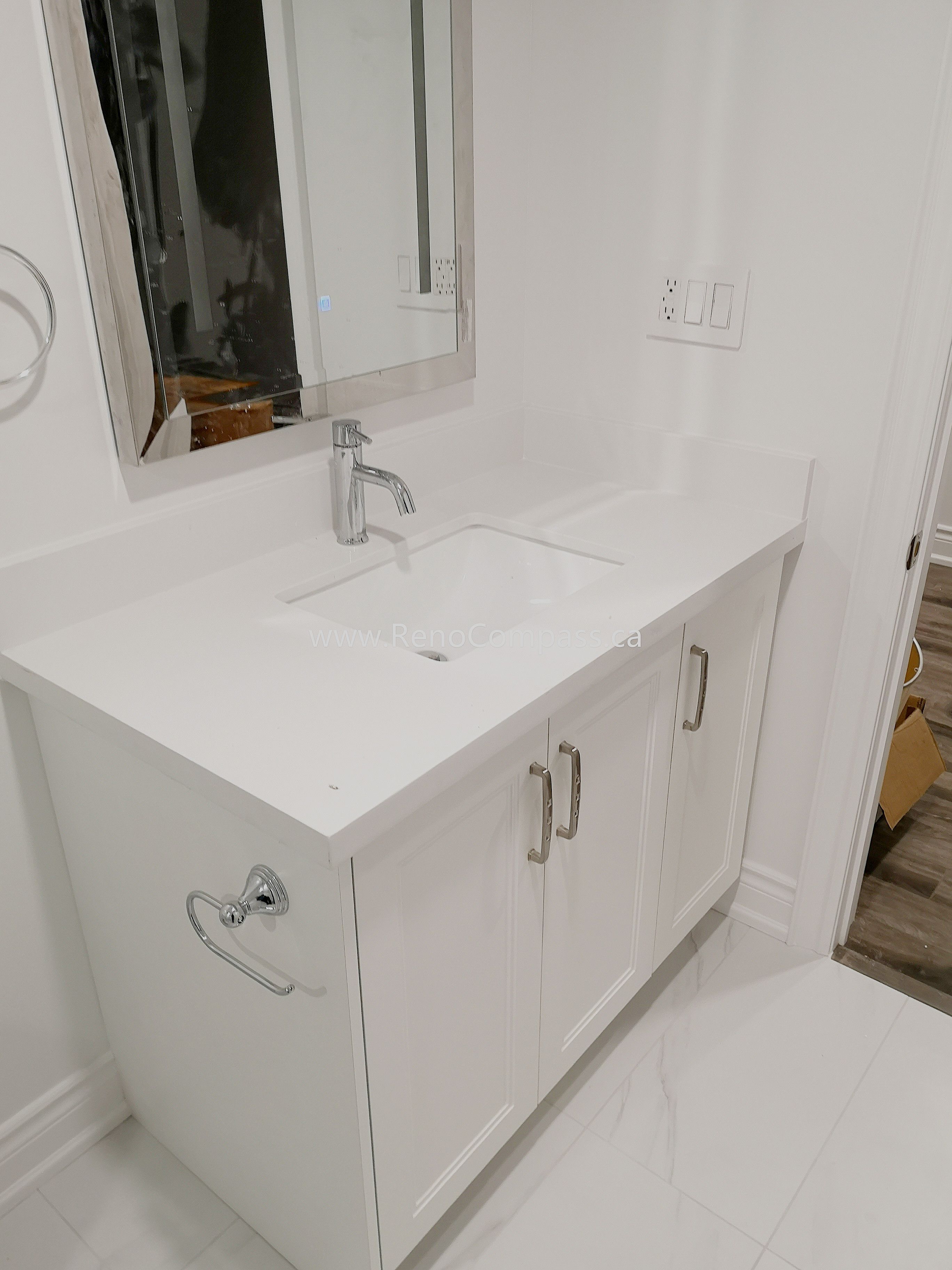 This Vaughan basement renovation features a modern bathroom vanity installation with a sleek white quartz countertop, under-mount sink, and contemporary chrome faucet. The clean white cabinetry provides both storage and a minimalist aesthetic, perfectly matching the bright and modern basement design. Completed by Reno Compass Inc., this basement bathroom upgrade enhances functionality while maintaining a stylish and cohesive interior renovation finish.