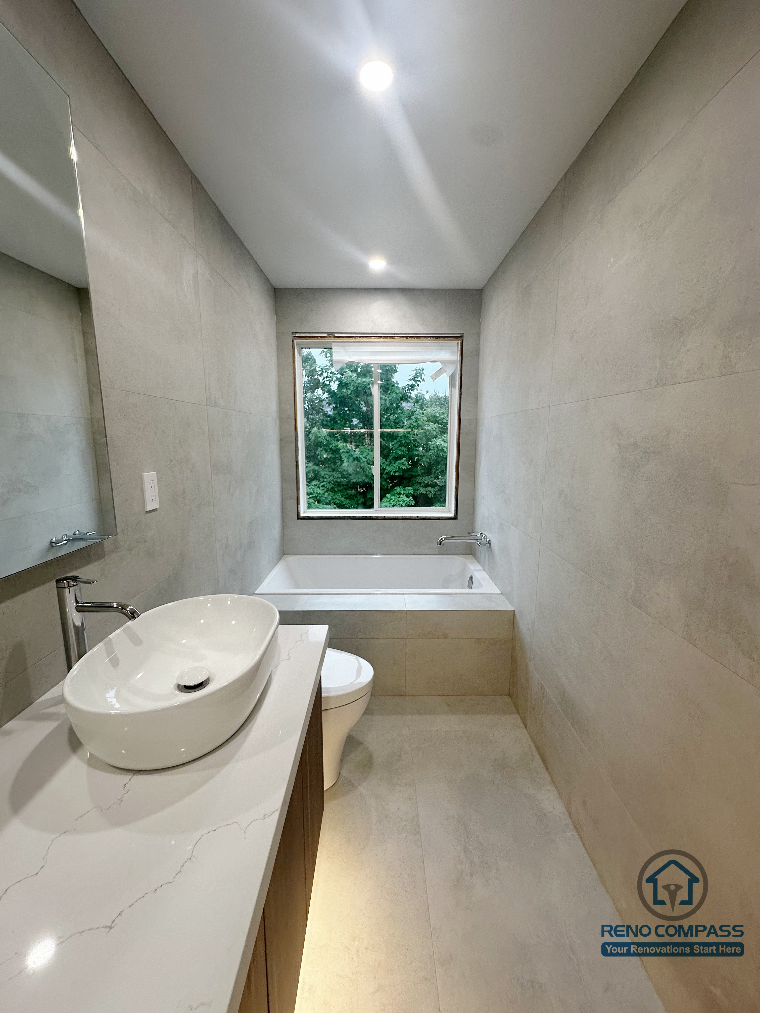 This Oakville bathroom renovation showcases a clean, linear layout featuring a vessel sink vanity, a quartz countertop, and a built-in soaking tub positioned beneath a large window. Large-format neutral wall tiles and recessed ceiling lighting create a calm, spa-inspired atmosphere, while the compact layout maximizes both comfort and functionality. Completed by Reno Compass, this bathroom is designed for everyday use with a focus on durability, light, and timeless design.