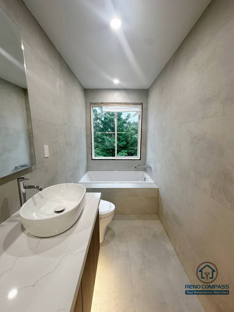 This Oakville bathroom renovation showcases a clean, linear layout featuring a vessel sink vanity, a quartz countertop, and a built-in soaking tub positioned beneath a large window. Large-format neutral wall tiles and recessed ceiling lighting create a calm, spa-inspired atmosphere, while the compact layout maximizes both comfort and functionality. Completed by Reno Compass, this bathroom is designed for everyday use with a focus on durability, light, and timeless design.