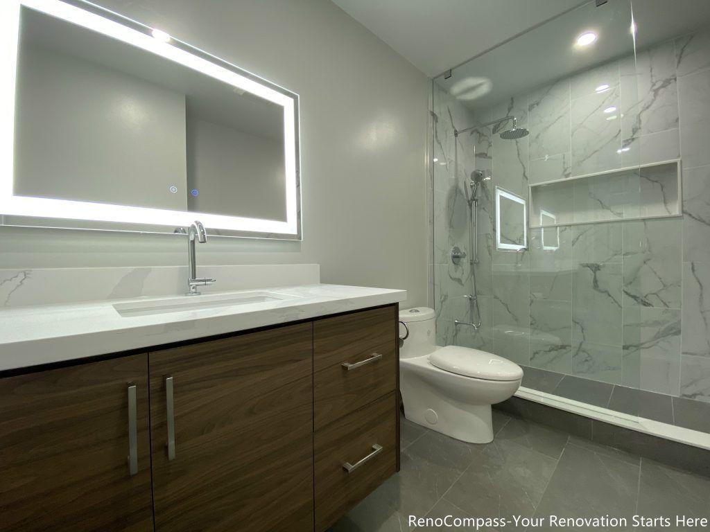 This Oakville bathroom renovation showcases a modern walk-in glass shower with marble-look wall tiles, built-in shower niche, and sleek frameless enclosure for a clean contemporary design. The space also features a custom wood vanity with quartz countertop, LED backlit mirror, and upgraded fixtures that enhance both functionality and visual appeal. Completed by a professional Toronto renovation company, this bathroom upgrade focuses on maximizing brightness, storage, and comfort while delivering a stylish and durable modern bathroom space.