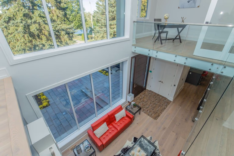 Modern open-concept living room in a 5000 sqft Toronto home renovation featuring double-height ceilings, glass railing mezzanine, wide plank hardwood flooring, contemporary fireplace wall with built-in TV, and large sliding doors leading to a backyard deck. The space combines natural light, clean architectural lines, and bold red leather sofas to create a bright and sophisticated interior. Renovated by Reno Compass Inc., a professional Toronto renovation company specializing in large-scale custom home interior transformations.