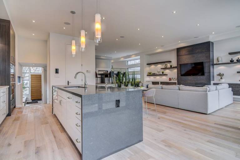 Modern open-concept kitchen and living room renovation in Toronto featuring a large quartz waterfall island, pendant lighting, light wood flooring, and custom built-in wall unit with fireplace and TV. The spacious layout seamlessly connects cooking, dining, and living areas, creating a bright and functional luxury home environment. Designed and completed by a professional Toronto renovation company, this space reflects modern comfort and upscale residential design.