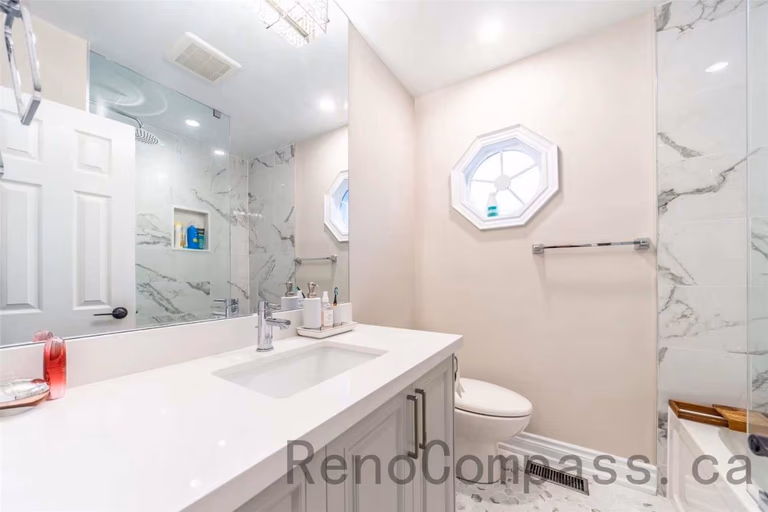 This modern bathroom is part of a North York interior renovation, featuring a clean and functional layout designed for daily comfort. The space includes a custom vanity with quartz countertop, an undermount sink, and sleek hardware for a refined look. Large mirrors and recessed ceiling lights enhance brightness, while marble-style wall tiles and a glass shower enclosure add a contemporary finish. Thoughtful material selection and efficient space planning reflect the quality and attention to detail expected from professional Toronto interior renovation projects.