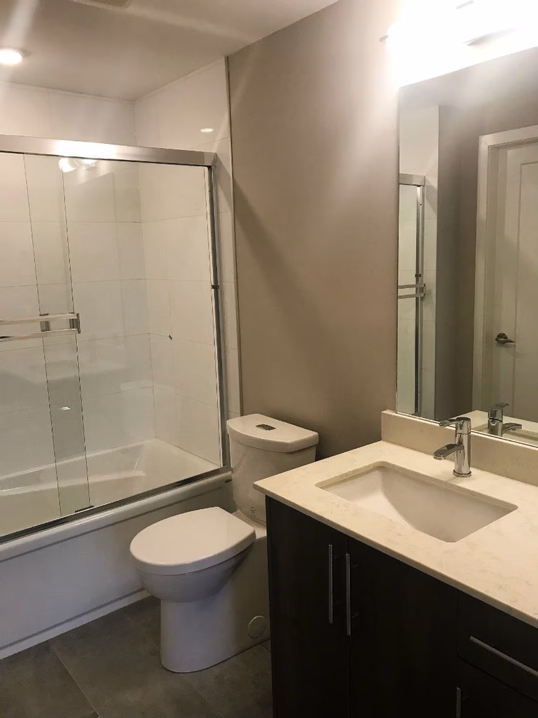 This Oakville condo bathroom renovation features a modern bathtub with frameless glass shower enclosure, sleek vanity with integrated sink, and contemporary tile flooring. The neutral color palette and upgraded lighting enhance brightness and functionality, creating a clean and comfortable everyday bathroom space in this professionally renovated condominium.
