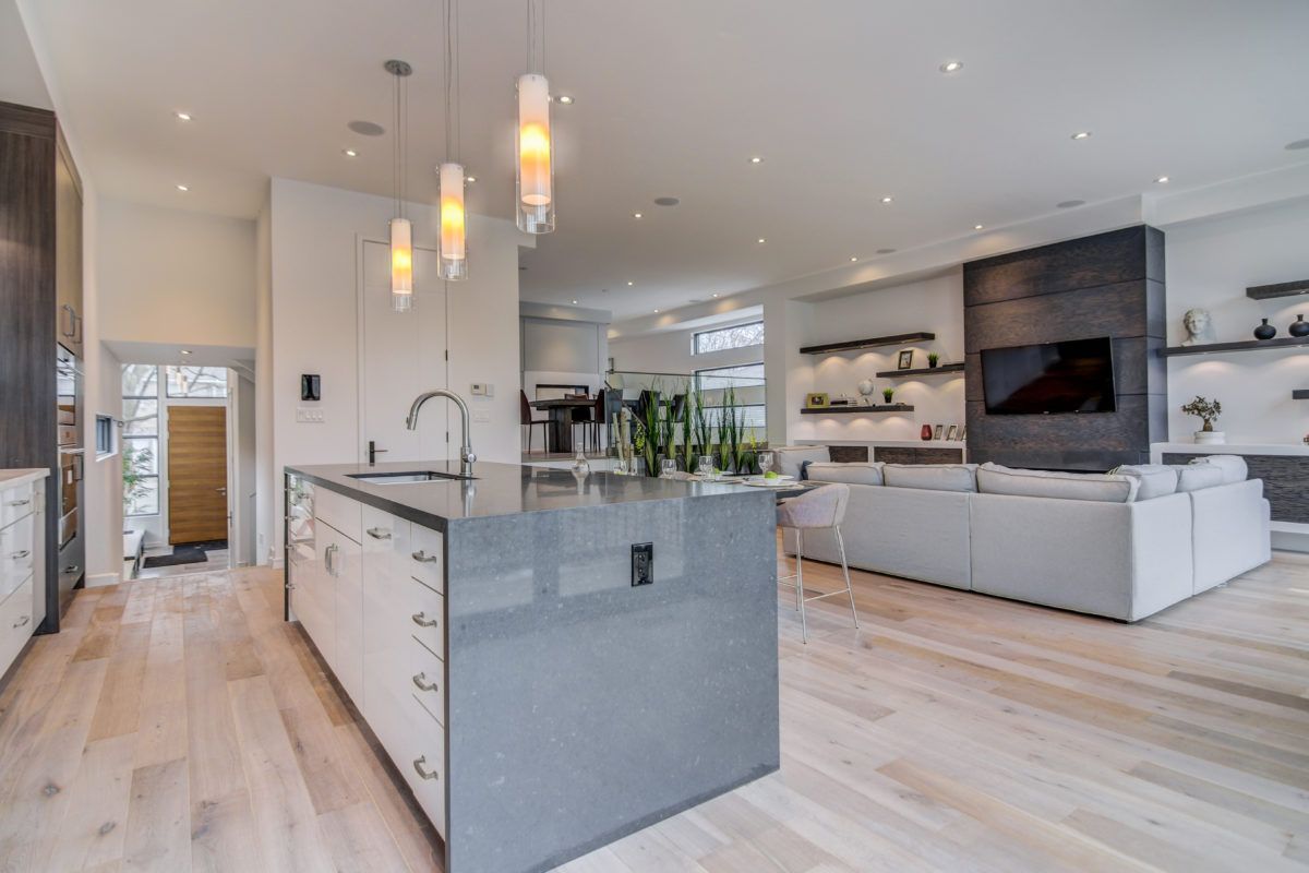 Modern open-concept kitchen and living room renovation in Toronto featuring a large quartz waterfall island, pendant lighting, light wood flooring, and custom built-in wall unit with fireplace and TV. The spacious layout seamlessly connects cooking, dining, and living areas, creating a bright and functional luxury home environment. Designed and completed by a professional Toronto renovation company, this space reflects modern comfort and upscale residential design.