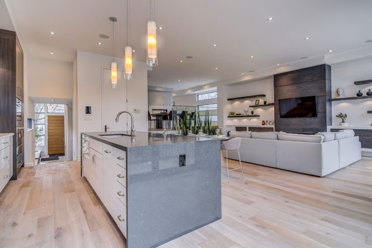 Modern open-concept kitchen and living room renovation in Toronto featuring a large quartz waterfall island, pendant lighting, light wood flooring, and custom built-in wall unit with fireplace and TV. The spacious layout seamlessly connects cooking, dining, and living areas, creating a bright and functional luxury home environment. Designed and completed by a professional Toronto renovation company, this space reflects modern comfort and upscale residential design.