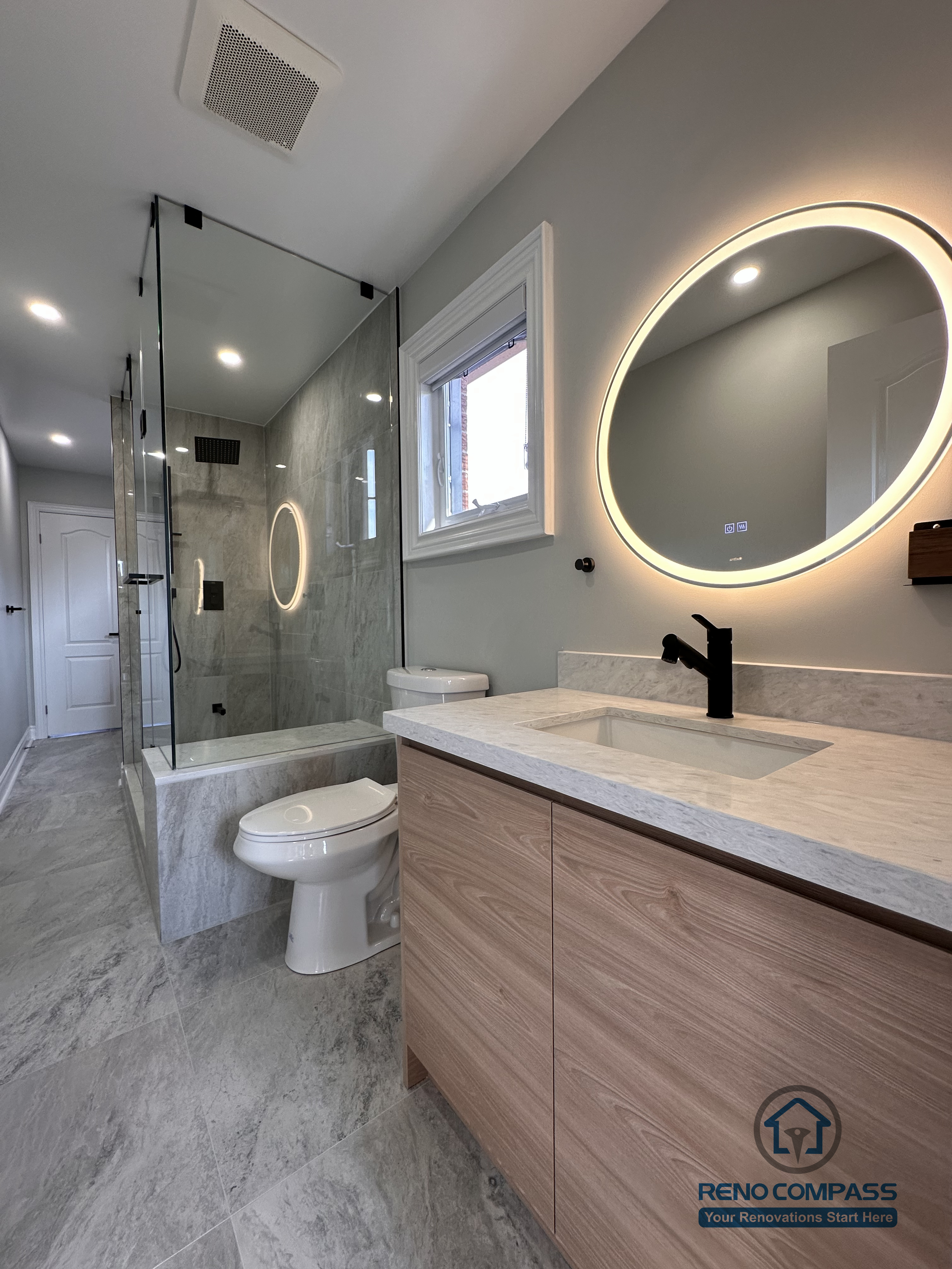 This Markham bathroom renovation features a custom wood-tone vanity with a quartz countertop, round LED backlit mirror, and matte black fixtures. The layout combines a glass-enclosed shower and bathtub with a practical vanity and toilet arrangement, creating a clean and well-balanced space. Designed and built by a professional Toronto renovation company, Reno Compass Inc., this bathroom emphasizes functionality, durability, and timeless design.