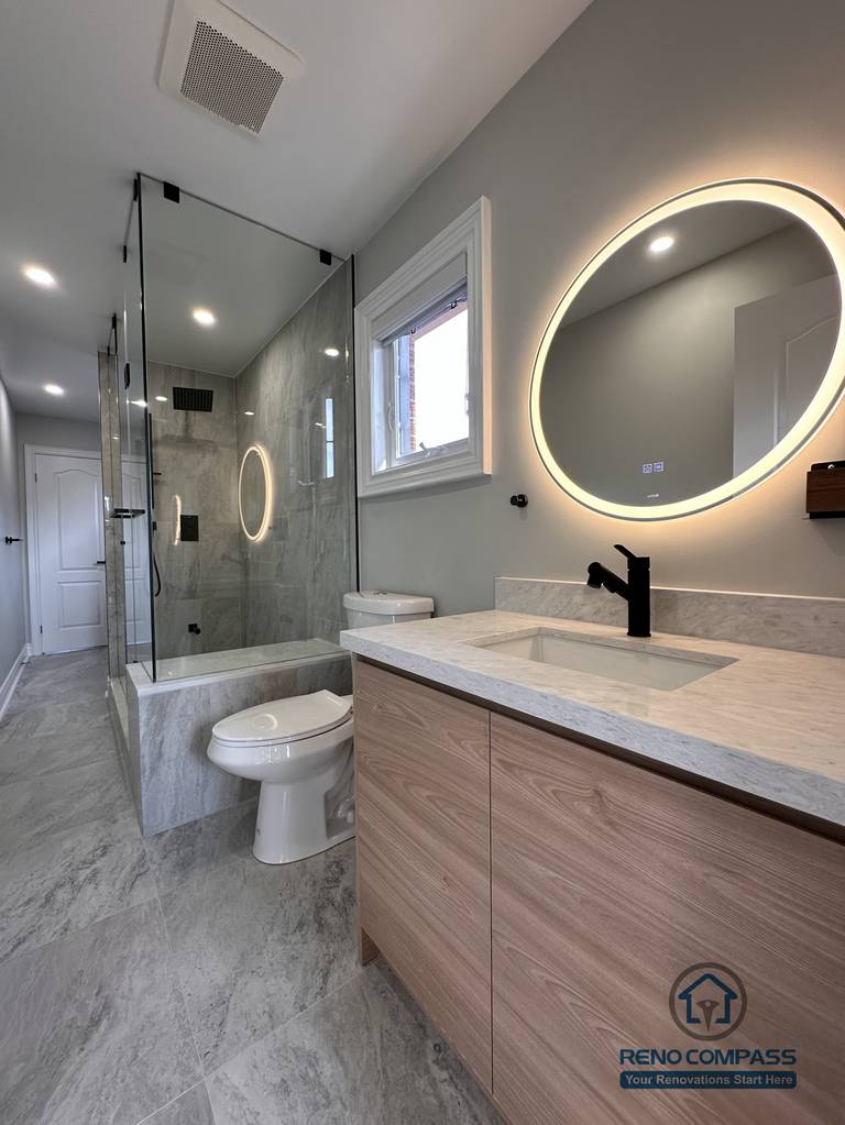 This Markham bathroom renovation features a custom wood-tone vanity with a quartz countertop, round LED backlit mirror, and matte black fixtures. The layout combines a glass-enclosed shower and bathtub with a practical vanity and toilet arrangement, creating a clean and well-balanced space. Designed and built by a professional Toronto renovation company, Reno Compass Inc., this bathroom emphasizes functionality, durability, and timeless design.