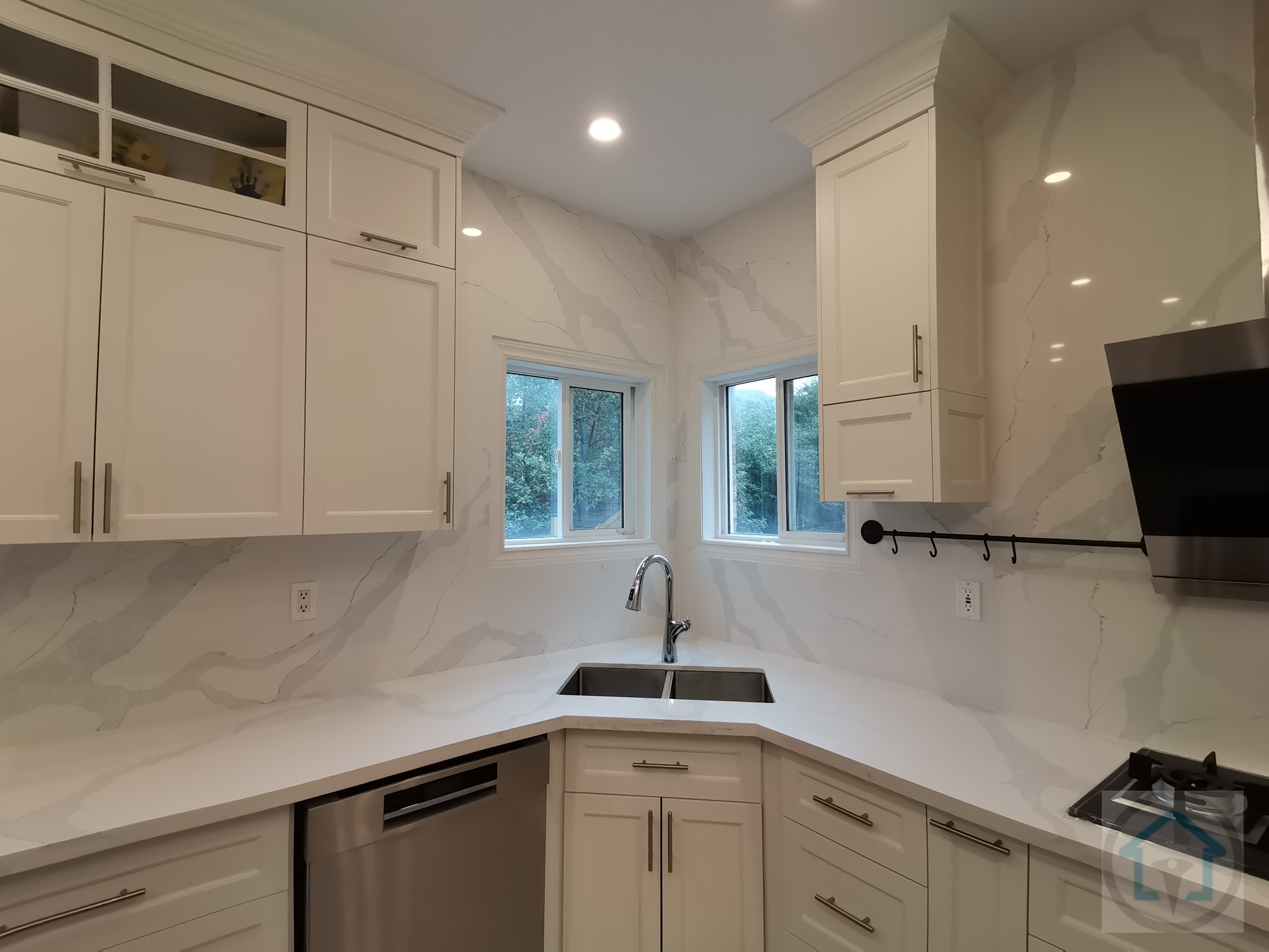 This kitchen renovation highlights a functional corner sink layout framed by dual windows, maximizing natural light and workspace efficiency. Full-height marble-look backsplash panels extend seamlessly from countertop to ceiling, creating a clean, continuous visual surface that elevates the overall design. Custom white cabinetry with integrated storage surrounds the corner configuration, while a stainless steel undermount sink and modern faucet enhance everyday usability. The thoughtful placement of cabinetry, lighting, and appliances ensures balanced proportions and smooth workflow. Completed by a professional Toronto renovation company, this kitchen renovation demonstrates how detailed planning and refined material selection can transform underused corners into elegant, high-performance spaces.