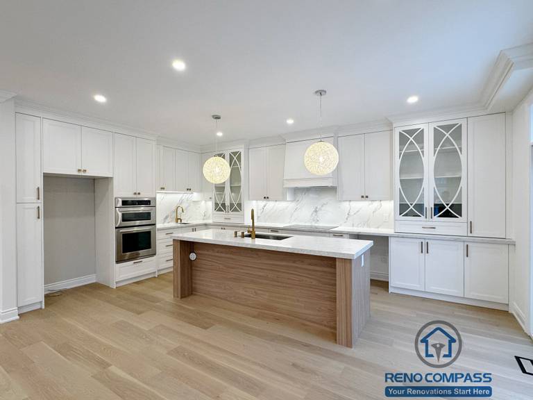Custom white kitchen completed as part of a Vaughan interior renovation, featuring a large waterfall island with wood paneling, quartz countertops, marble-look backsplash, glass-front cabinets, built-in appliances, and modern pendant lighting. The clean layout and premium finishes highlight the craftsmanship of a professional Toronto renovation company.
