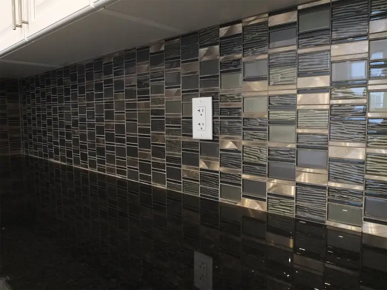 This Kleinburg interior renovation highlights a close-up view of the modern mosaic tile backsplash installed behind the kitchen countertop. The combination of glass and textured tiles in dark tones creates a stylish contrast against the sleek black countertop and white cabinetry. Precision tile installation enhances both durability and visual depth, adding a sophisticated finishing detail to the contemporary kitchen design.
