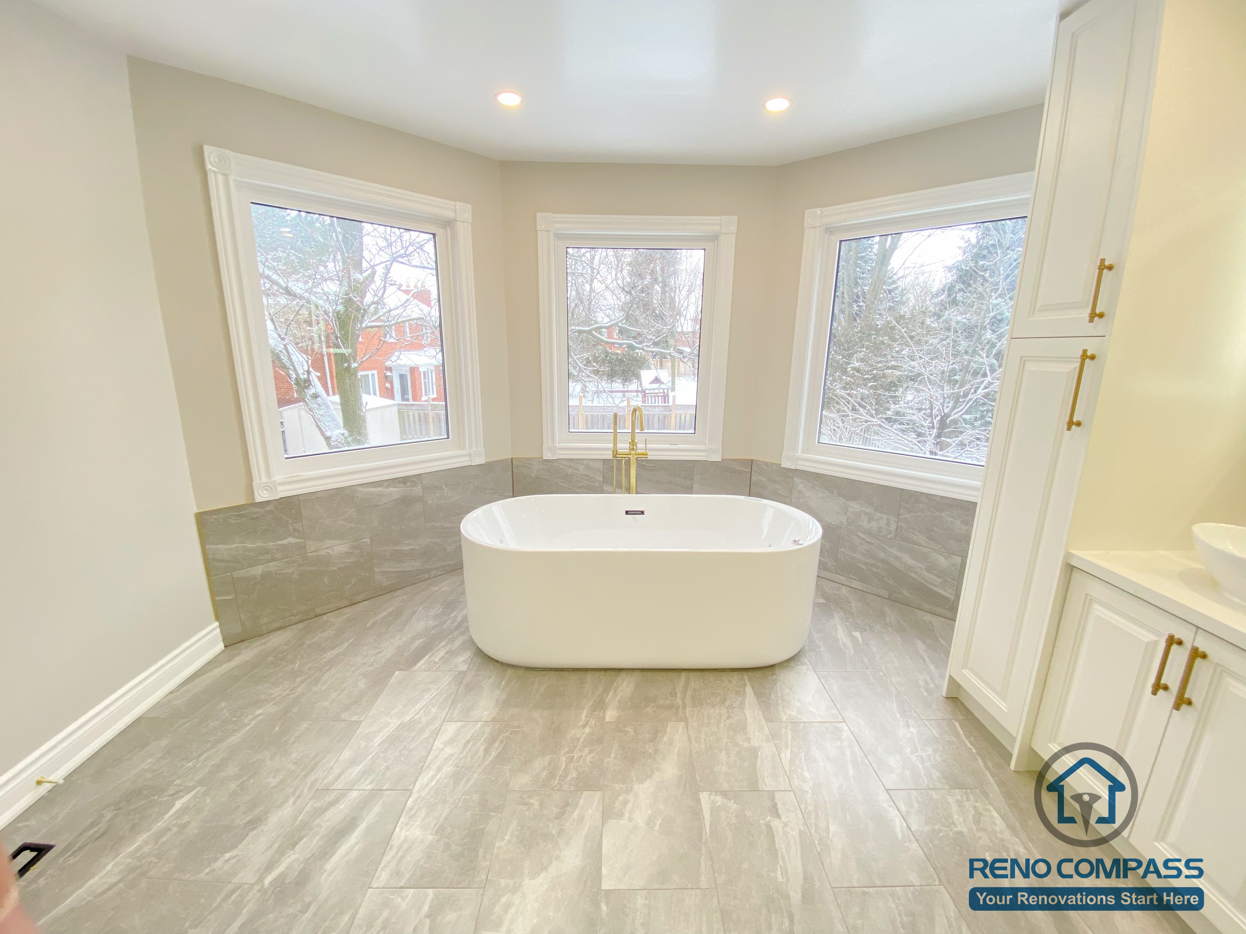 This Richmond Hill bathroom renovation highlights a modern freestanding bathtub positioned in front of large bay windows, creating a bright and relaxing spa-like atmosphere. The space features large-format grey floor tiles, marble-style wall finishes, recessed ceiling lighting, and gold-finish tub fixtures. The clean layout and natural light enhance comfort, elegance, and functionality, showcasing a thoughtfully designed luxury bathroom renovation in Richmond Hill.