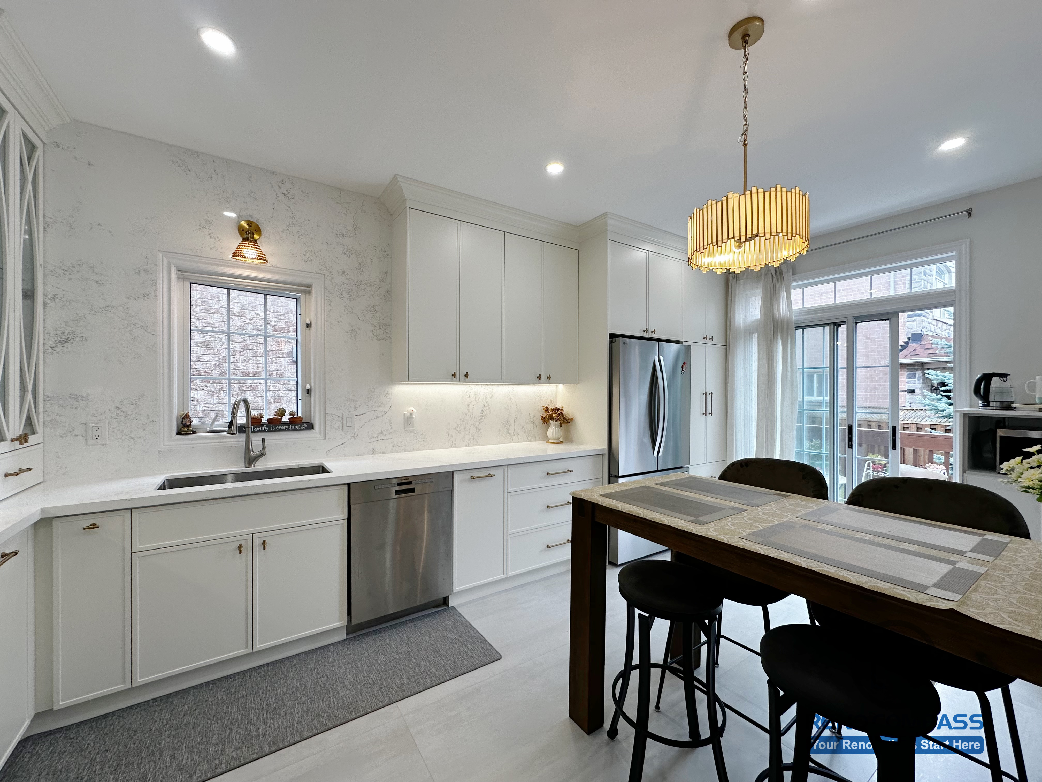This Markham kitchen renovation showcases a bright and well-balanced layout with custom white cabinetry, quartz countertops, and a functional dining island. A statement chandelier adds warmth and visual focus above the seating area, while integrated lighting enhances both task efficiency and overall ambiance. Large windows and patio doors allow natural light to flow through the space, creating a kitchen designed for everyday comfort, family dining, and seamless indoor-outdoor living.