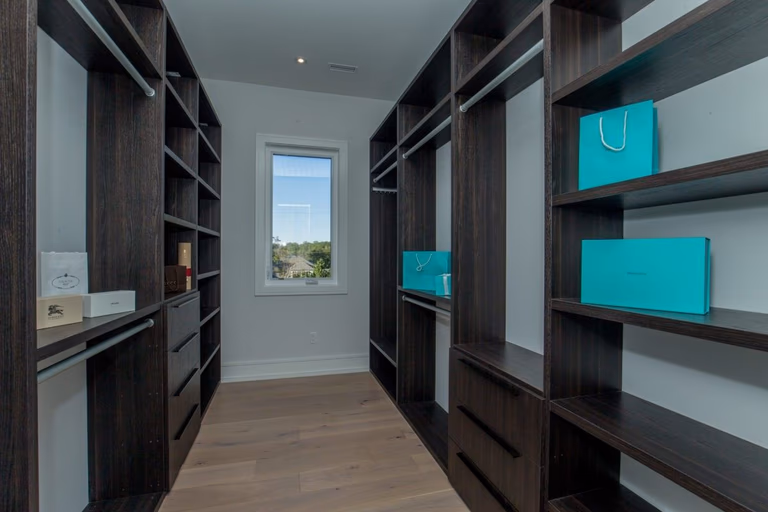 Luxury walk-in closet renovation in a 5000 sqft Toronto custom home featuring dark wood built-in shelving, full-height wardrobes, integrated drawers, and open display storage for organized dressing space. This modern closet design maximizes storage efficiency while maintaining a clean, high-end interior aesthetic with natural lighting and hardwood flooring. Completed by Reno Compass Inc., a professional Toronto renovation company specializing in custom interior renovations and luxury home upgrades.
