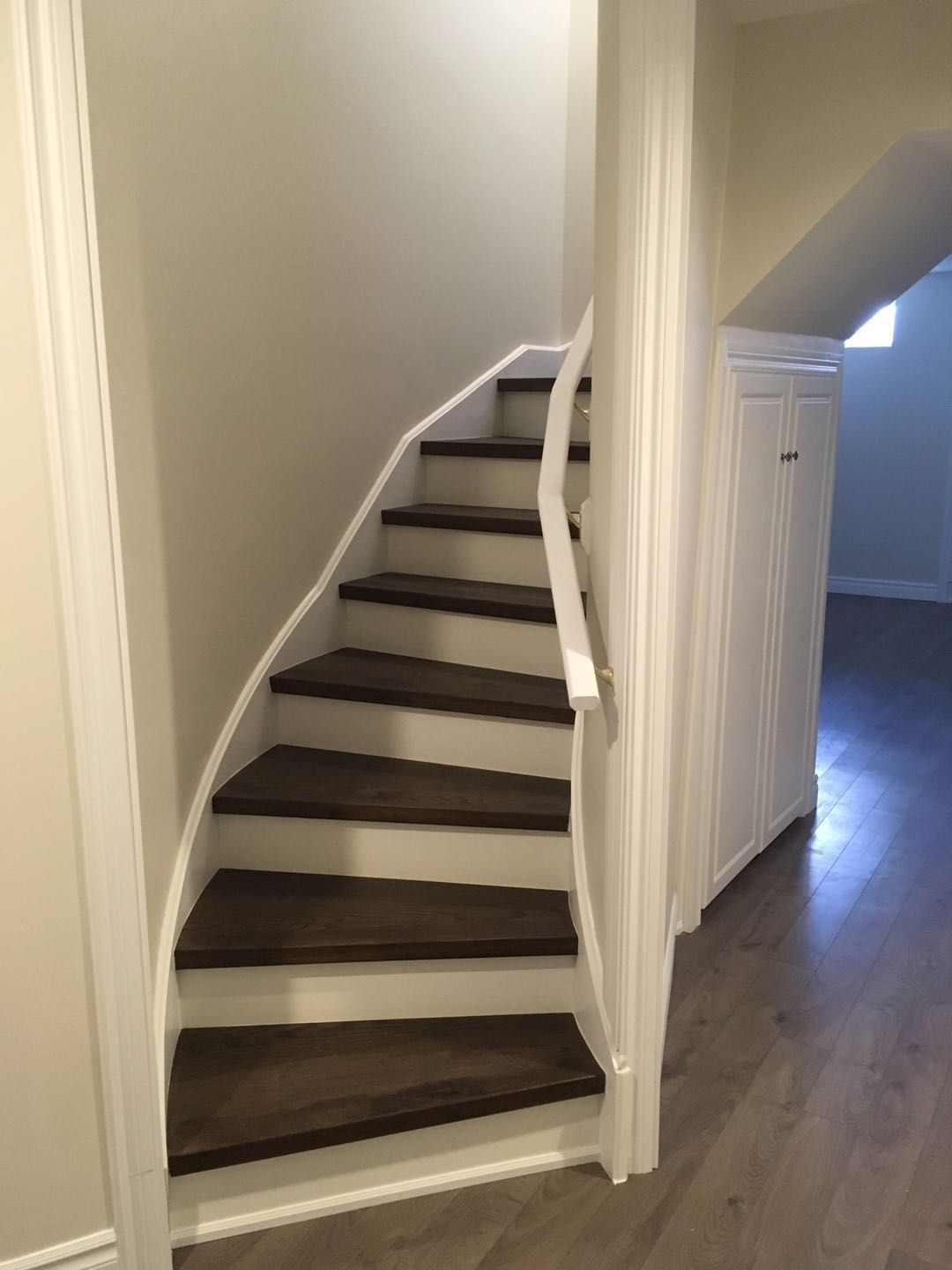 This Toronto interior renovation showcases a custom curved staircase featuring rich dark wood treads paired with clean white risers and railings. The design maximizes space efficiency while adding architectural interest, seamlessly connecting levels with a modern yet timeless aesthetic. The surrounding neutral tones and hardwood flooring enhance the overall warmth and flow of the home.