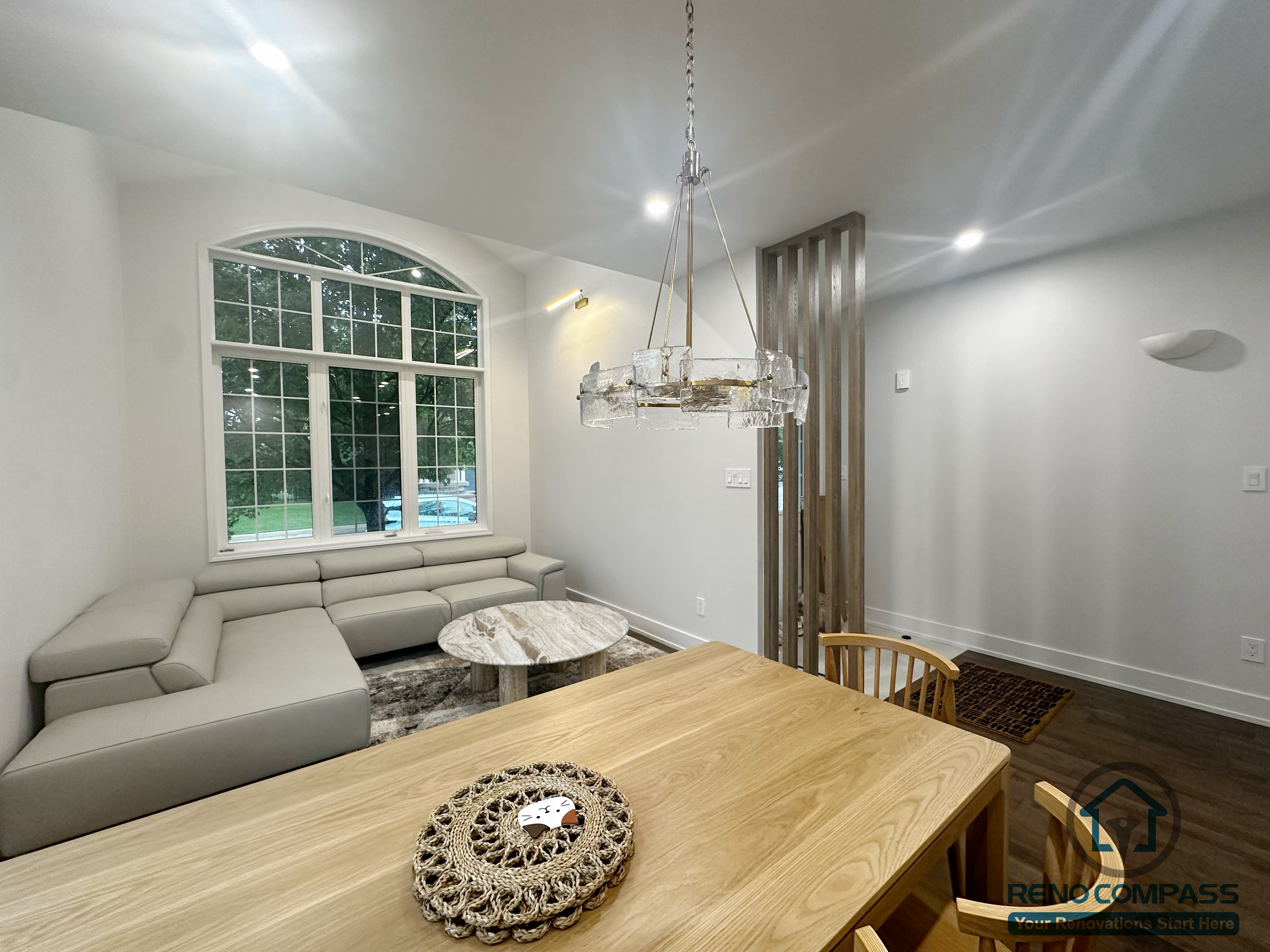 This Oakville interior renovation captures the relationship between the dining area and adjacent living space from a connected perspective. The solid wood dining table anchors the foreground, while the living area beyond is framed by a large arched window that brings in natural light. A neutral sectional sofa and marble coffee table create a calm, comfortable seating zone. The wood slat divider subtly defines circulation without closing off the space, maintaining openness and visual flow. The layout emphasizes proportion, light, and everyday functionality, supporting long-term use in a modern family home.