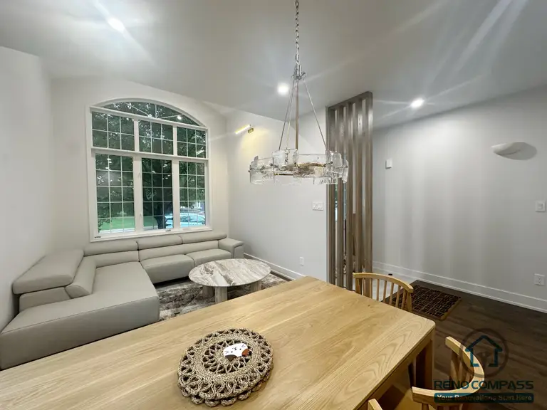 This Oakville interior renovation captures the relationship between the dining area and adjacent living space from a connected perspective. The solid wood dining table anchors the foreground, while the living area beyond is framed by a large arched window that brings in natural light. A neutral sectional sofa and marble coffee table create a calm, comfortable seating zone. The wood slat divider subtly defines circulation without closing off the space, maintaining openness and visual flow. The layout emphasizes proportion, light, and everyday functionality, supporting long-term use in a modern family home.