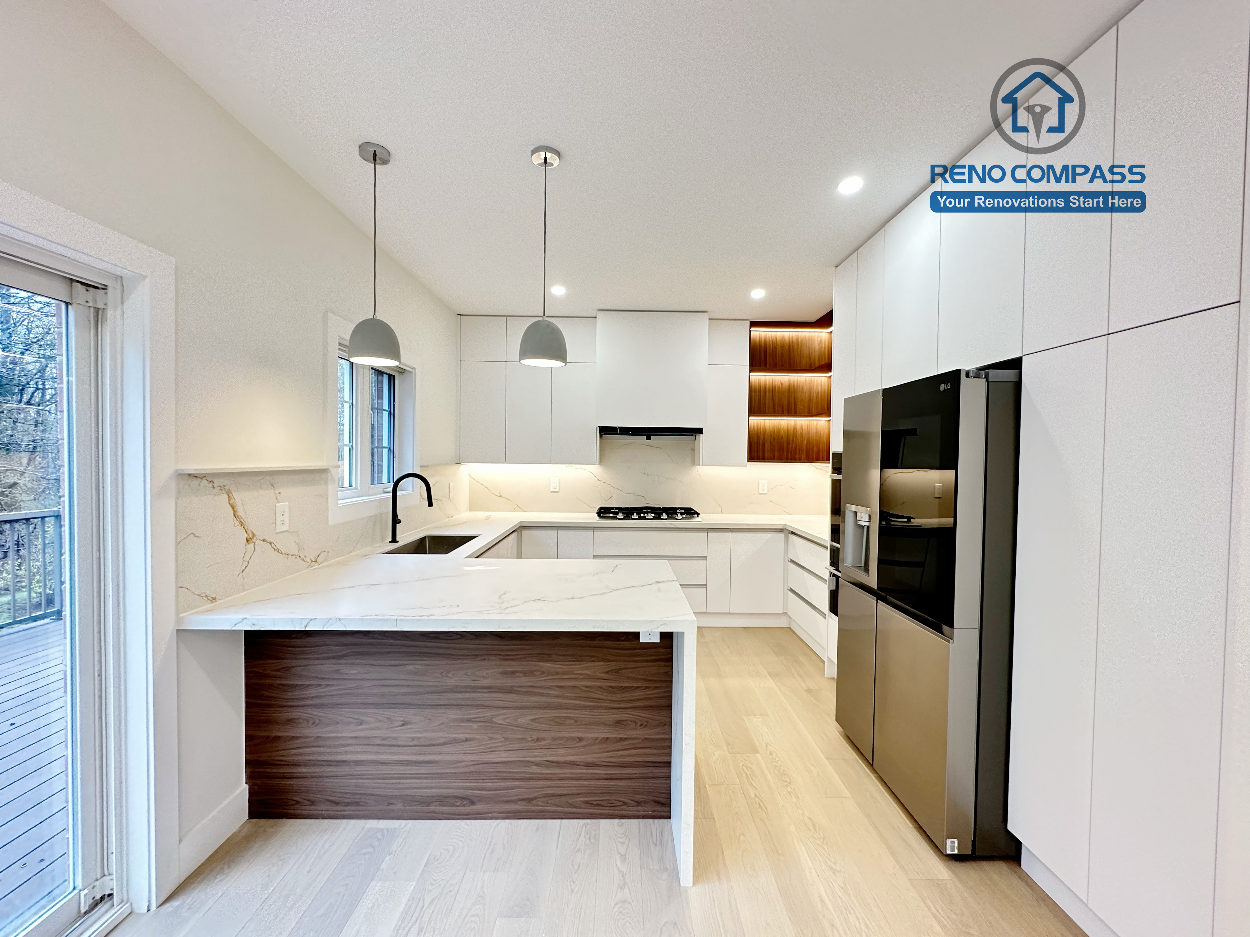 This image presents a modern kitchen renovation in Richmond Hill featuring full-height custom white cabinetry, a quartz waterfall island with wood panel detailing, and integrated stainless steel appliances. The design includes a built-in gas cooktop, minimalist pendant lights, recessed ceiling lighting, and light hardwood flooring for a clean, contemporary look. Large windows and patio access enhance natural light and openness. Completed by a professional Toronto Renovation Company, this kitchen renovation reflects refined craftsmanship, efficient layout planning, and modern family-focused design.