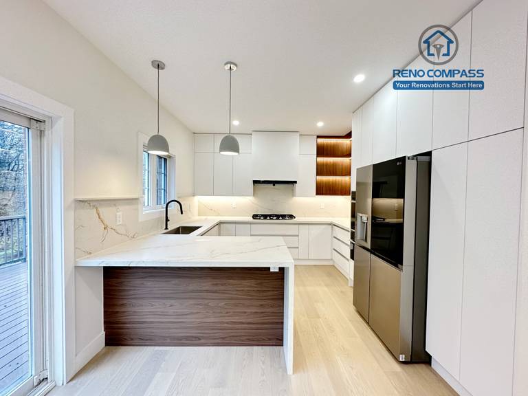 This image presents a modern kitchen renovation in Richmond Hill featuring full-height custom white cabinetry, a quartz waterfall island with wood panel detailing, and integrated stainless steel appliances. The design includes a built-in gas cooktop, minimalist pendant lights, recessed ceiling lighting, and light hardwood flooring for a clean, contemporary look. Large windows and patio access enhance natural light and openness. Completed by a professional Toronto Renovation Company, this kitchen renovation reflects refined craftsmanship, efficient layout planning, and modern family-focused design.