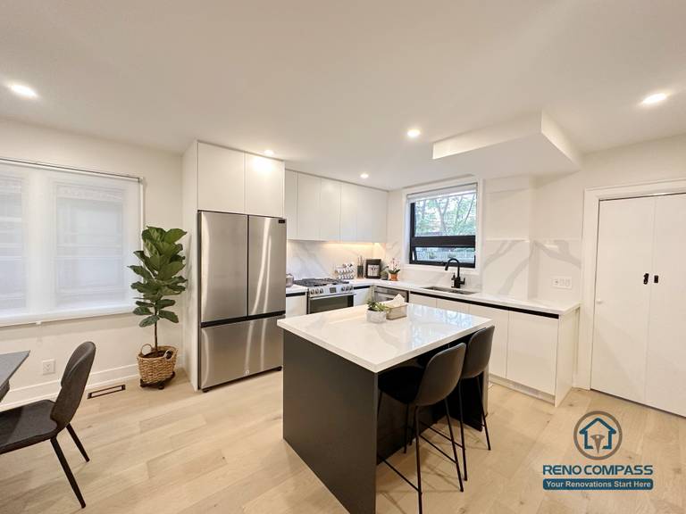 This Toronto interior renovation showcases a modern white kitchen featuring flat-panel cabinetry, a quartz waterfall island, stainless steel appliances, and recessed ceiling lighting. The clean lines and functional layout highlight contemporary kitchen design and quality workmanship delivered by a professional Toronto renovation company.