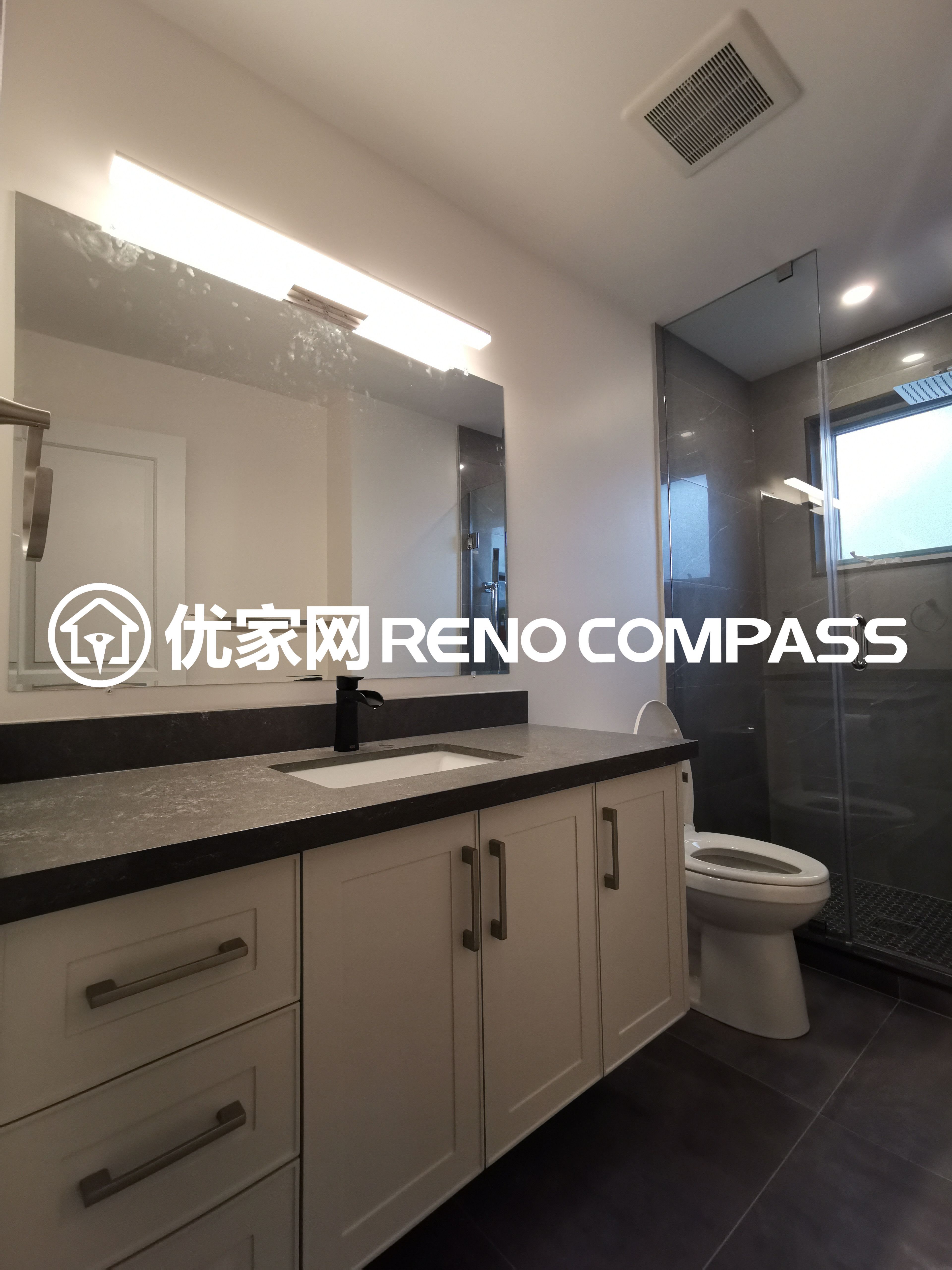 This image showcases a spacious bathroom completed as part of a North York interior renovation project. The design features a custom double-sink vanity with a quartz countertop, offering ample storage and functionality for daily use. A large mirror with integrated LED lighting enhances brightness, while the glass-enclosed shower adds a modern and streamlined look. Neutral cabinetry and dark floor tiles create a balanced contrast, reflecting quality craftsmanship and efficient space planning suited for contemporary Toronto homes.