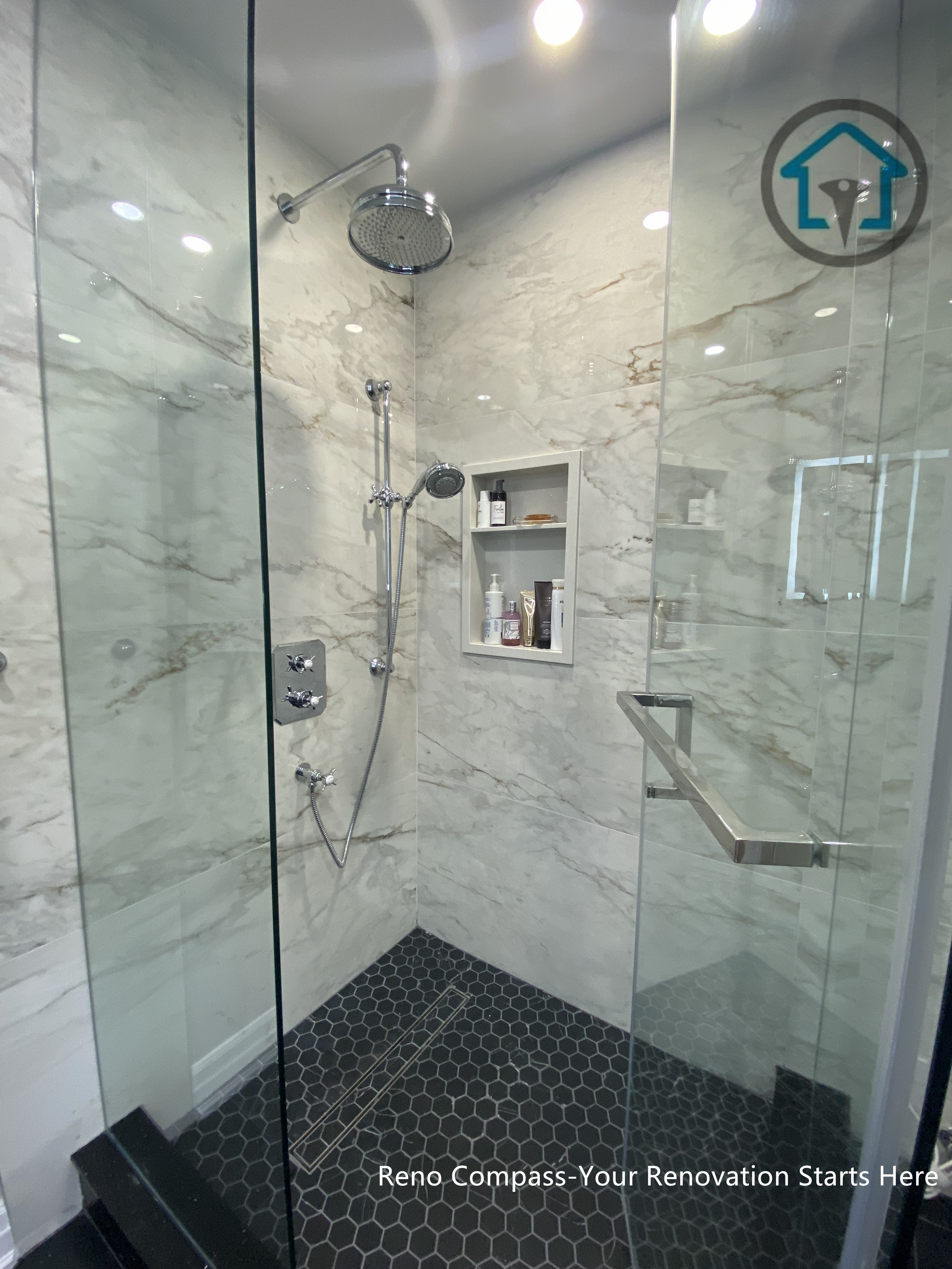 This Toronto bathroom renovation showcases a frameless glass shower paired with marble-look wall tiles and a dark hexagon mosaic shower base. The contrast between light stone surfaces and darker flooring defines the shower zone while maintaining a clean, modern appearance. Built-in shower niches provide practical storage without interrupting the visual flow, and the clear glass enclosure keeps the space feeling open and bright. The layout prioritizes comfort, durability, and ease of daily maintenance.