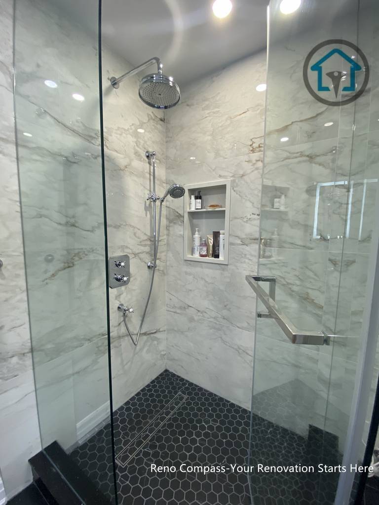 This Toronto bathroom renovation showcases a frameless glass shower paired with marble-look wall tiles and a dark hexagon mosaic shower base. The contrast between light stone surfaces and darker flooring defines the shower zone while maintaining a clean, modern appearance. Built-in shower niches provide practical storage without interrupting the visual flow, and the clear glass enclosure keeps the space feeling open and bright. The layout prioritizes comfort, durability, and ease of daily maintenance.