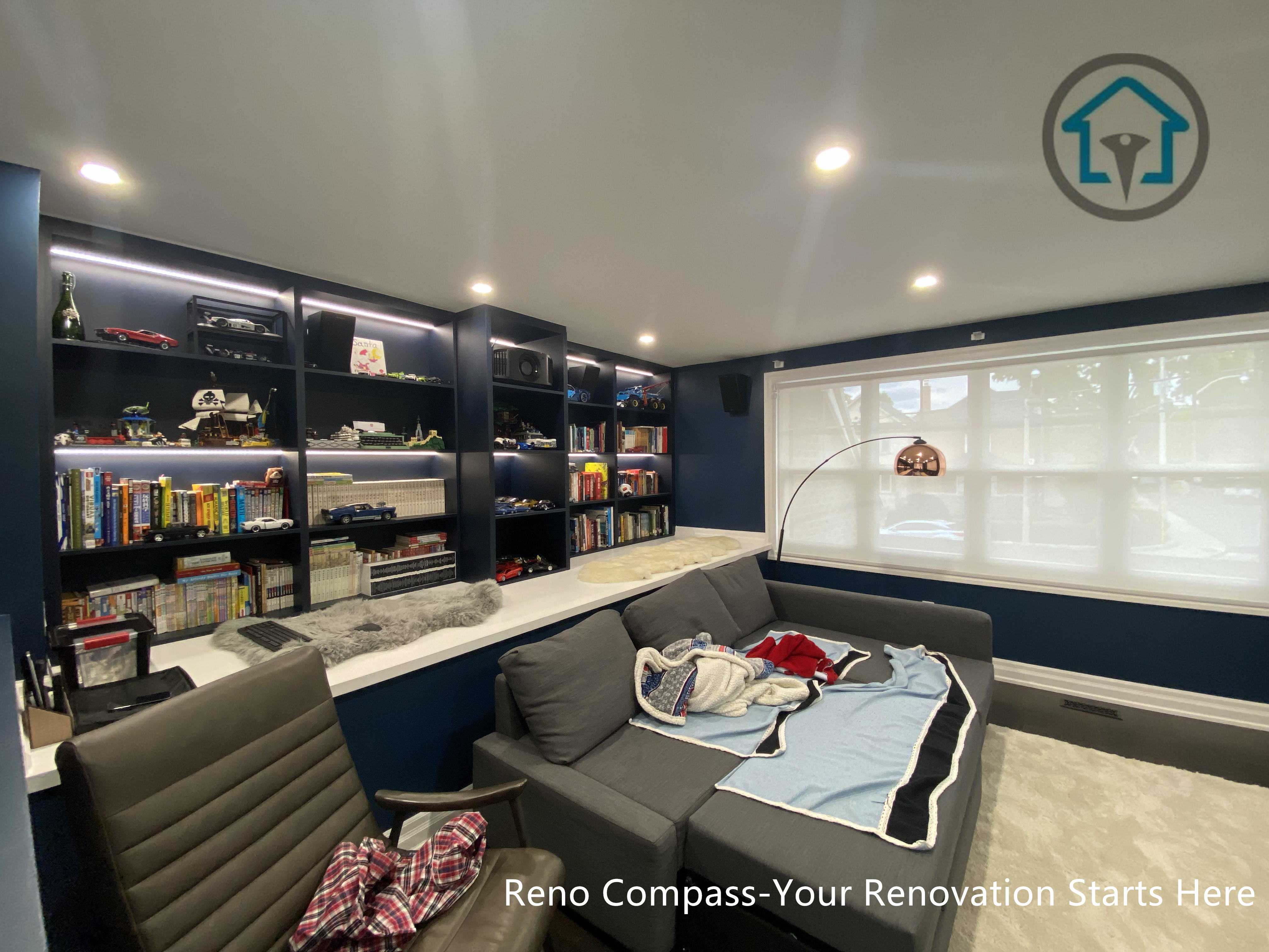 This image showcases a custom interior renovation project in Toronto, featuring a fully integrated built-in shelving and media wall designed for family leisure and everyday living. The dark-toned cabinetry provides generous storage while incorporating subtle accent lighting to enhance depth and visual balance. A comfortable seating area anchors the space, creating a flexible environment for reading, entertainment, and relaxation. The layout prioritizes functionality, lighting control, and long-term usability, reflecting a thoughtful approach to modern residential interior design.