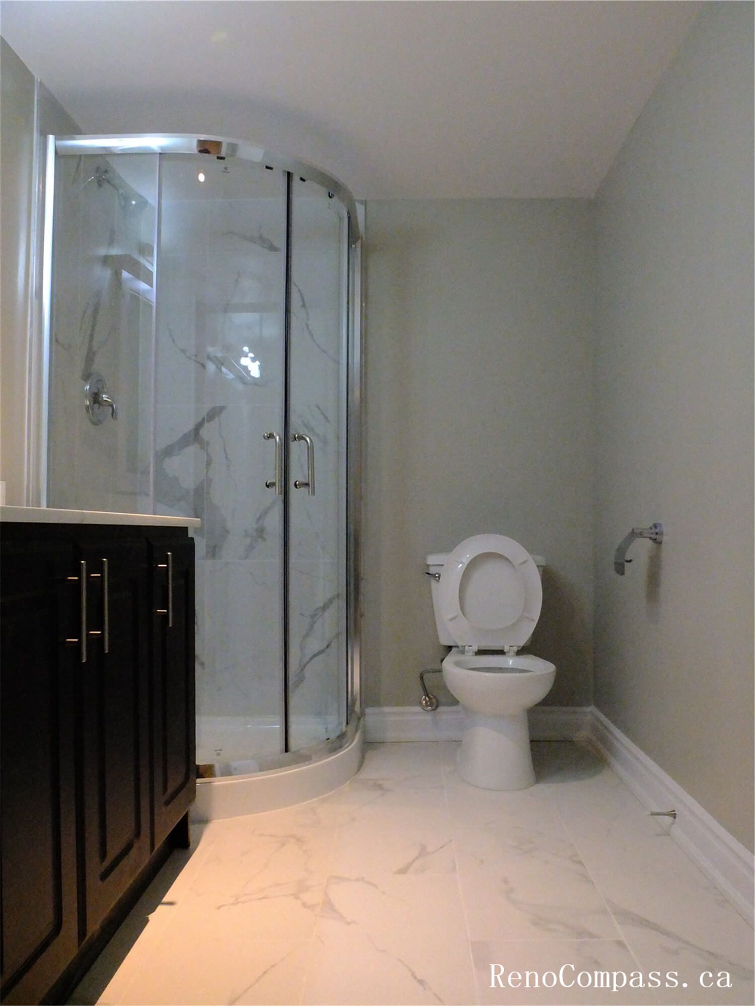 This Ajax basement renovation includes a modern bathroom featuring a curved glass shower enclosure with marble-style wall tiles, a sleek vanity with storage cabinetry, and bright porcelain floor tiles. The clean layout and contemporary finishes create a functional and stylish bathroom space for the finished basement. Completed by a professional Toronto Renovation Company, the design emphasizes comfort, durability, and efficient use of space in basement living areas.
