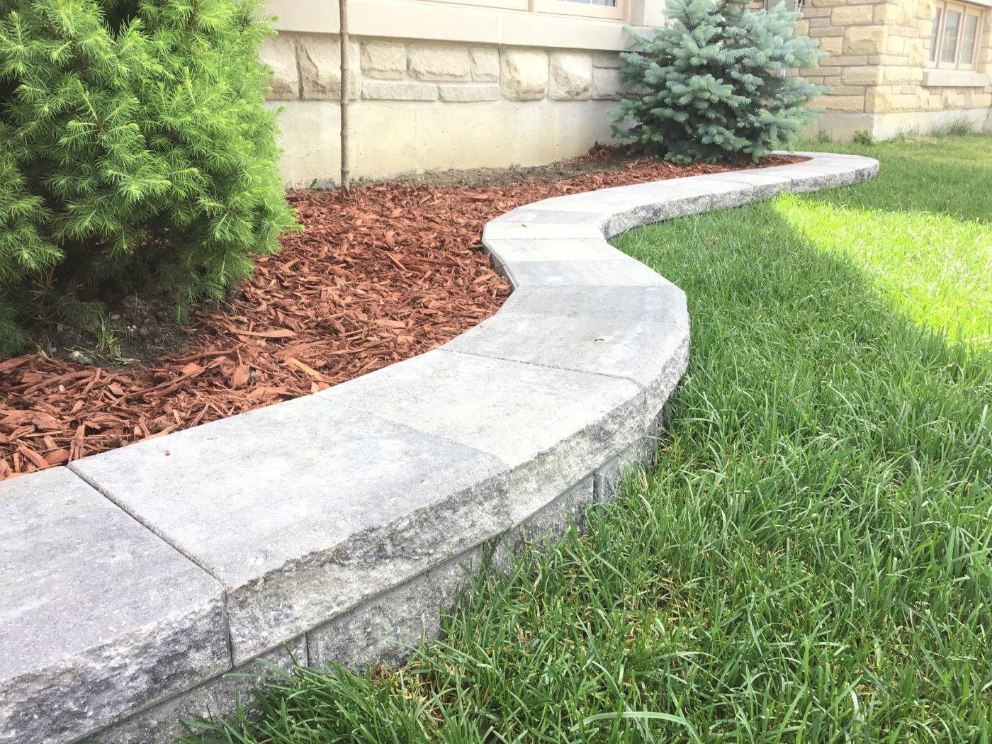 This Markham landscaping project showcases a curved stone retaining wall paired with a neatly finished mulch garden bed and evergreen shrubs. The smooth curved design adds visual flow to the landscape while improving soil retention and enhancing the overall curb appeal.