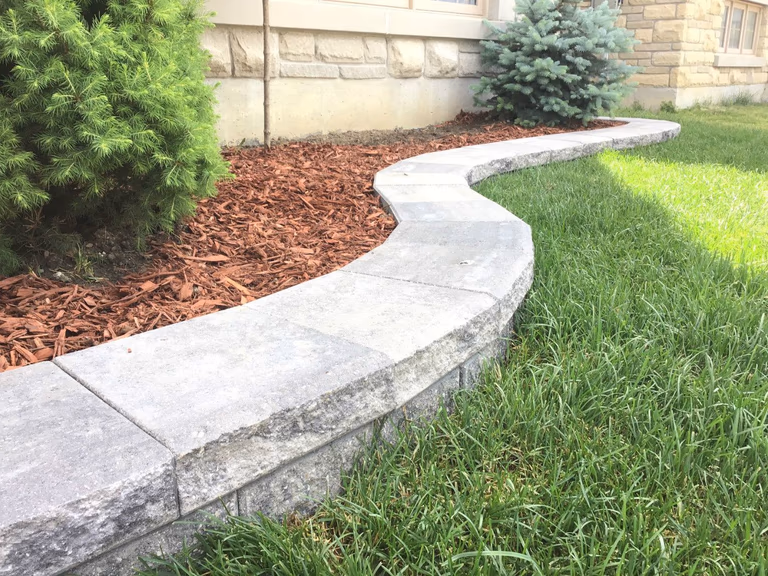 This Markham landscaping project showcases a curved stone retaining wall paired with a neatly finished mulch garden bed and evergreen shrubs. The smooth curved design adds visual flow to the landscape while improving soil retention and enhancing the overall curb appeal.